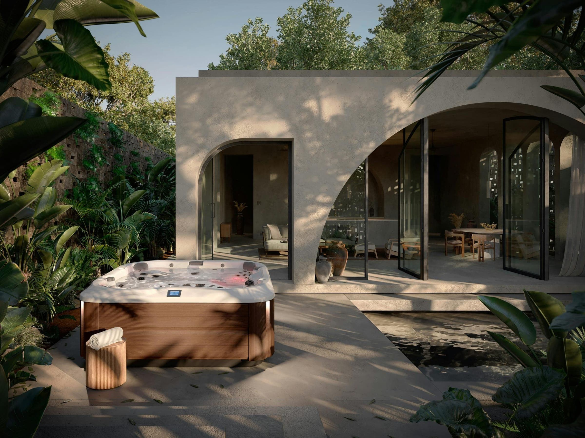 Spa de acrílico blanco veteado y gabinete café con acabado de madera en un patio de piedra, junto a un pabellón de estuco con grandes arcos y puertas de vidrio abiertas; alrededor, plantas tropicales, un espejo de agua y un banco redondo de madera con una toalla enrollada.