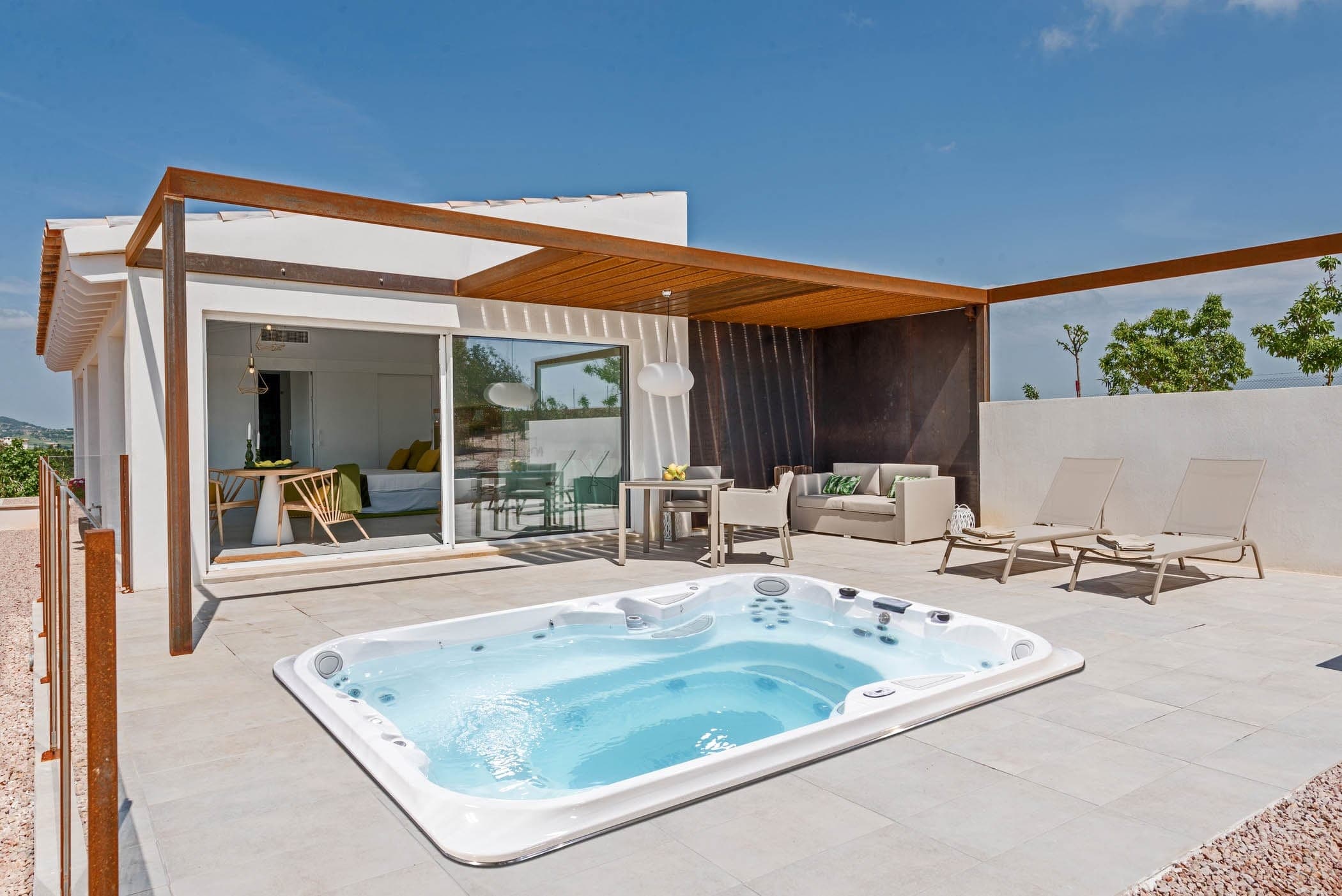 Swim spa blanco empotrado al ras en una terraza de piso pétreo claro, frente a una casa blanca con pérgola de acero oxidado y madera; alrededor, un comedor, una sala exterior y dos camastros bajo el cielo azul.