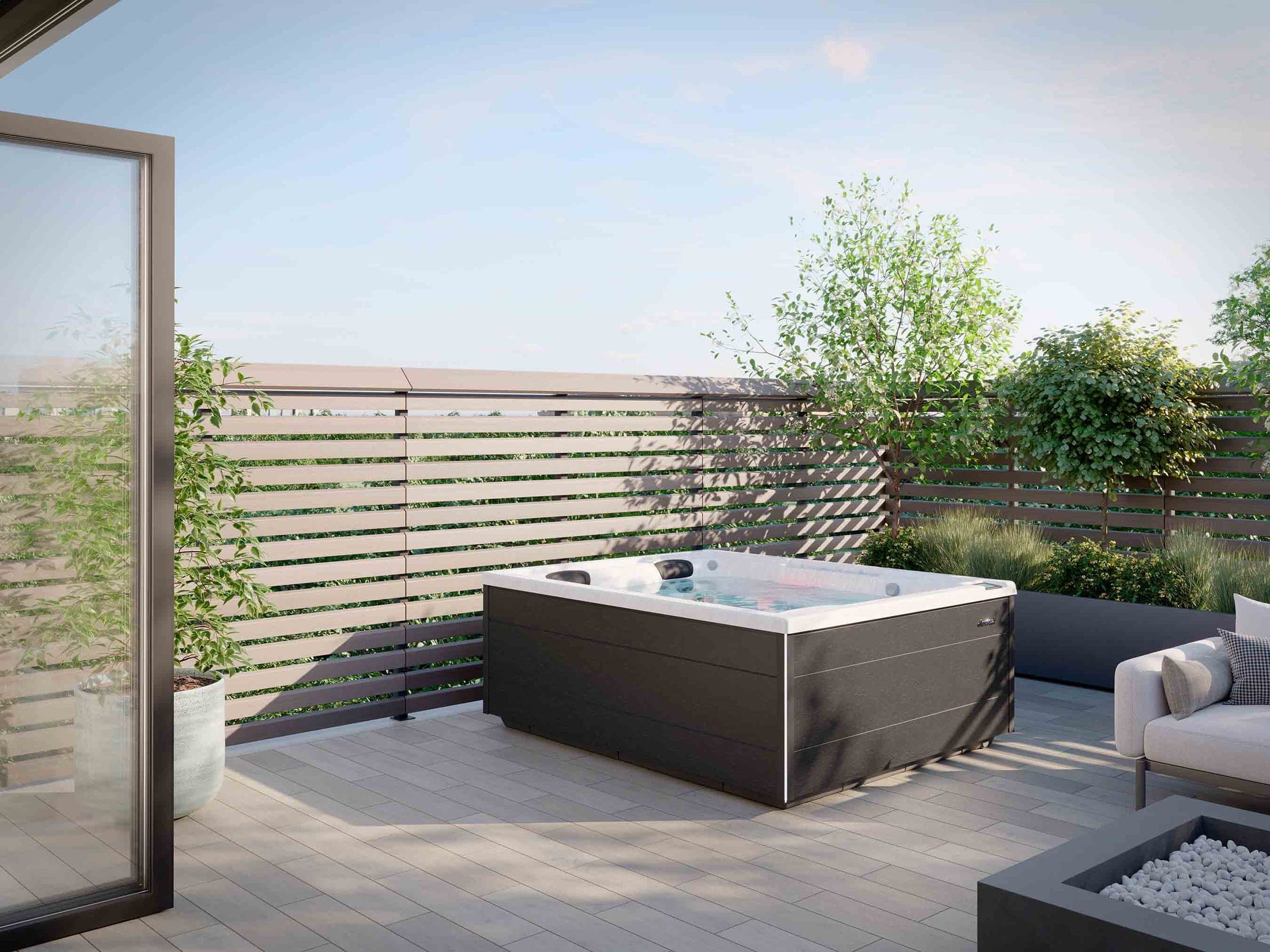 Spa rectangular de acrílico blanco y gabinete gris oscuro en una terraza de piso gris, con celosía de listones horizontales, jardineras, un sofá y un fogatero con piedras blancas; a la izquierda, una puerta de vidrio abierta.