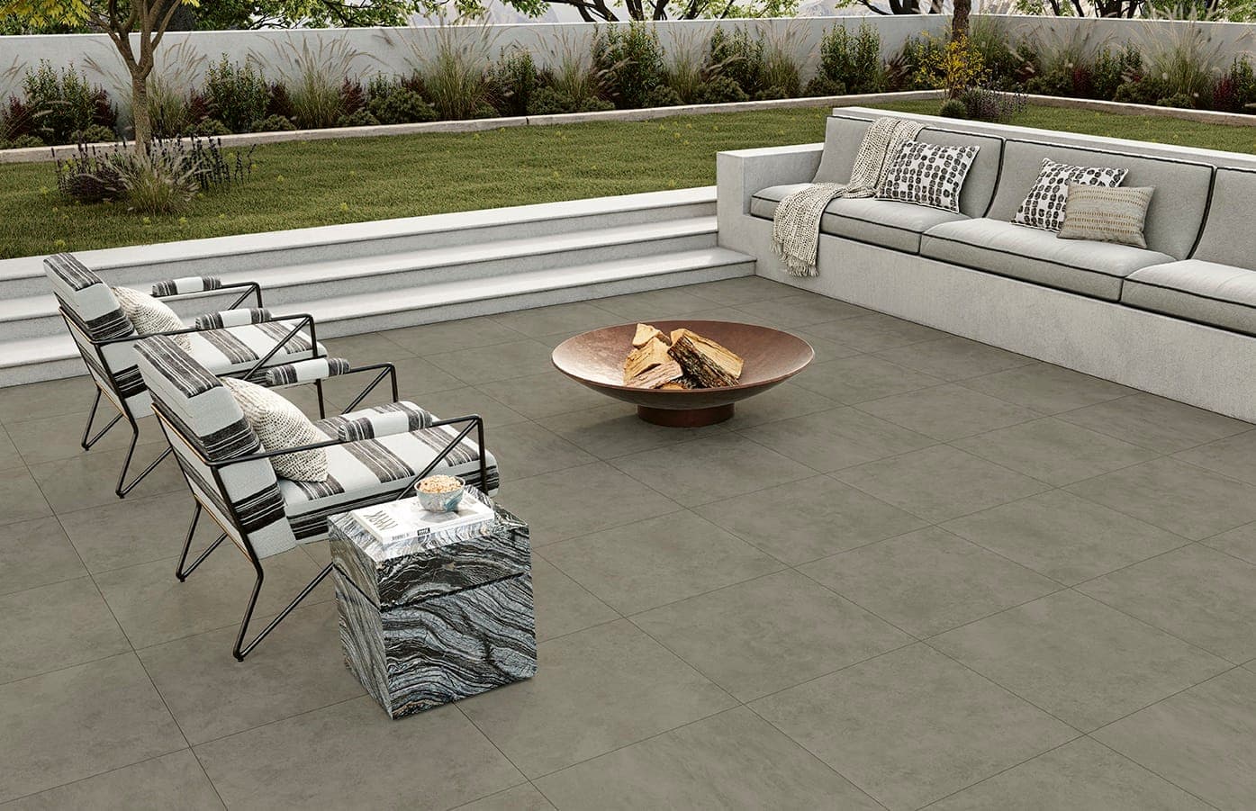Terraza con piso gris de piezas cuadradas, dos sillas de exterior con cojines a rayas, una mesa auxiliar de piedra veteada, un brasero de cobre con leña y una banca empotrada con cojines junto a escalones y jardín.