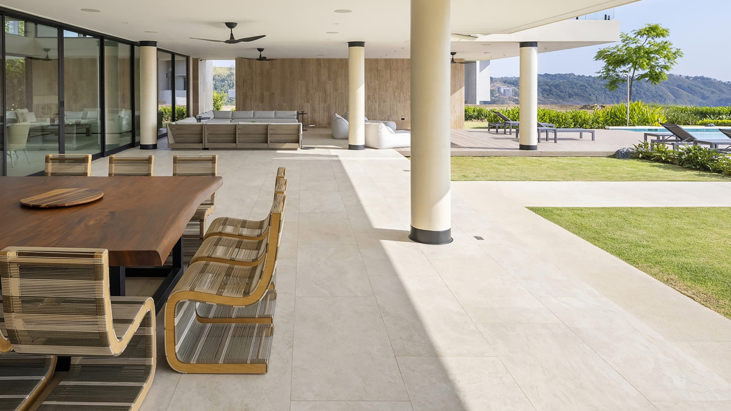 Terraza techada con piso claro de placas grandes que continúa en un andador junto al pasto; mesa de comedor de madera con sillas de cuerda, columnas blancas, una sala exterior al fondo y camastros junto a una alberca frente a cerros.