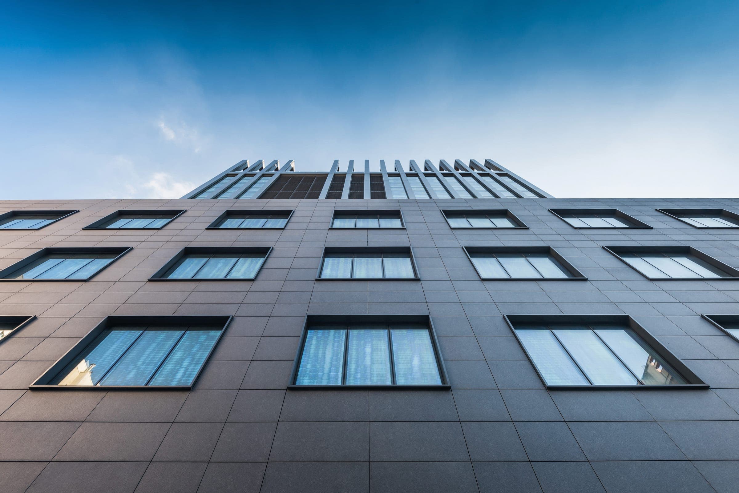 Fachada de un edificio vista desde abajo, revestida con placas rectangulares de porcelanato gris oscuro con juntas visibles, ventanas de marco oscuro y un remate de lamas verticales contra el cielo azul.