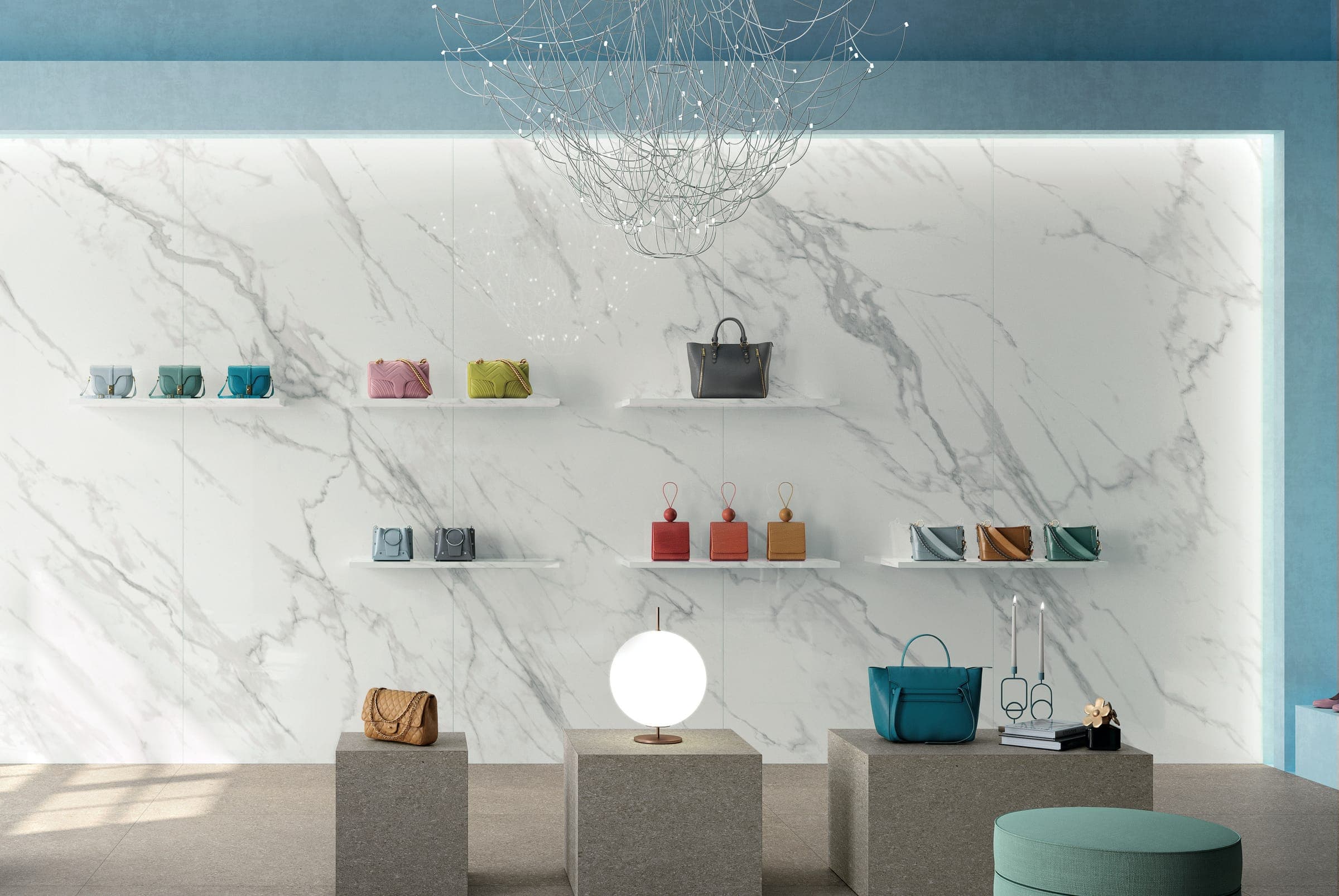 Tienda con muros azules y un muro de placas grandes efecto mármol blanco con vetas grises continuas; repisas con bolsas de mano de colores, una lámpara colgante de alambre, cubos de exhibición grises con una lámpara de globo y un puf verde agua.
