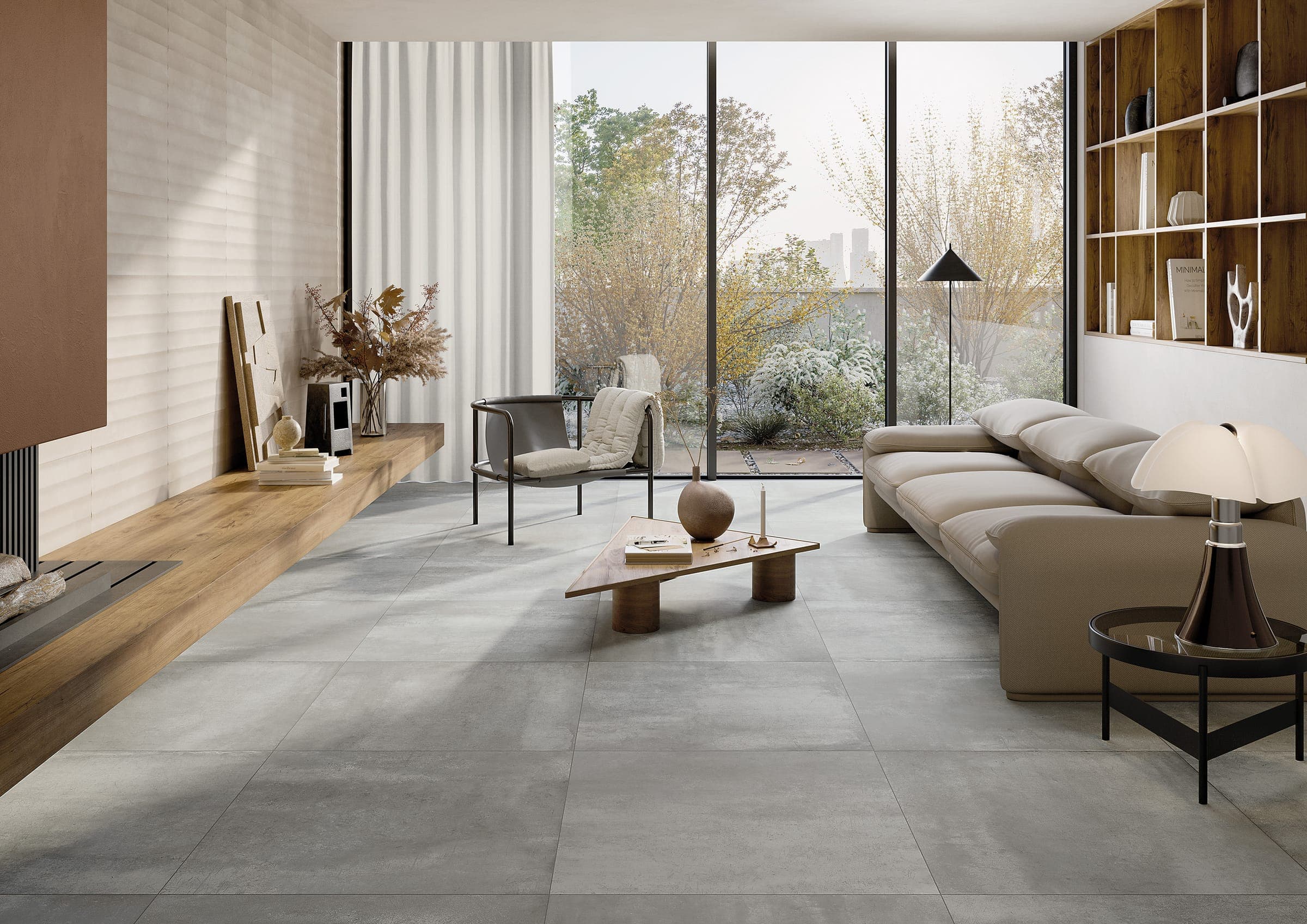 Sala con piso gris de placas grandes tipo cemento, un sofá beige, un sillón negro con una cobija, una mesa de centro triangular de madera, una banca de madera a lo largo de un muro claro con relieve horizontal, un librero de madera y un ventanal hacia un jardín.