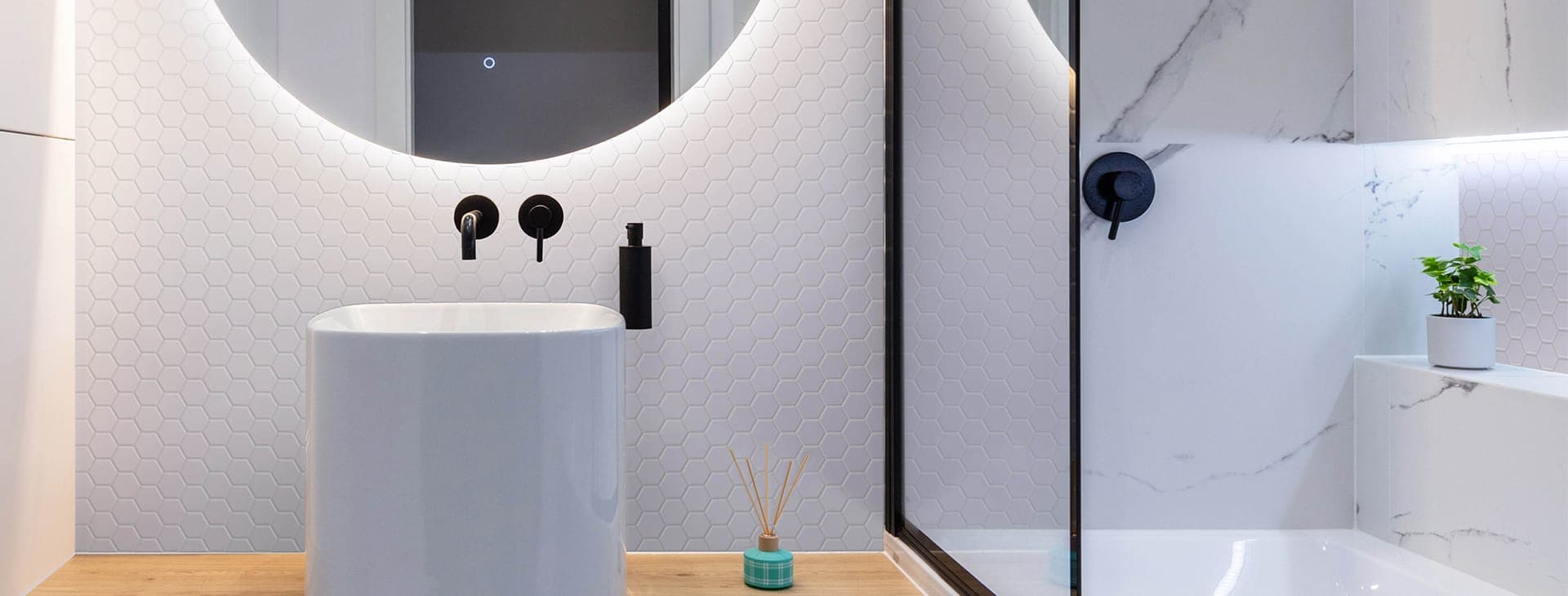 Baño con muro de mosaico hexagonal blanco mate, lavabo blanco sobre una superficie de madera, llaves negras empotradas, dispensador negro, espejo redondo iluminado y un difusor; a la derecha, un cancel de vidrio con perfil negro.