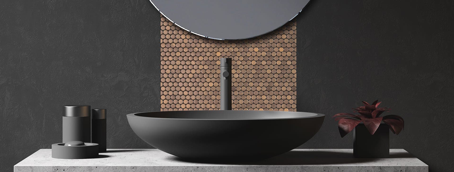 Muro gris oscuro con una franja vertical de mosaico circular tipo penny en tono cobrizo, detrás de un lavabo ovalado gris oscuro con llave alta y bajo un espejo redondo; a los lados, accesorios oscuros y una planta de hojas rojizas, sobre una cubierta de concreto.