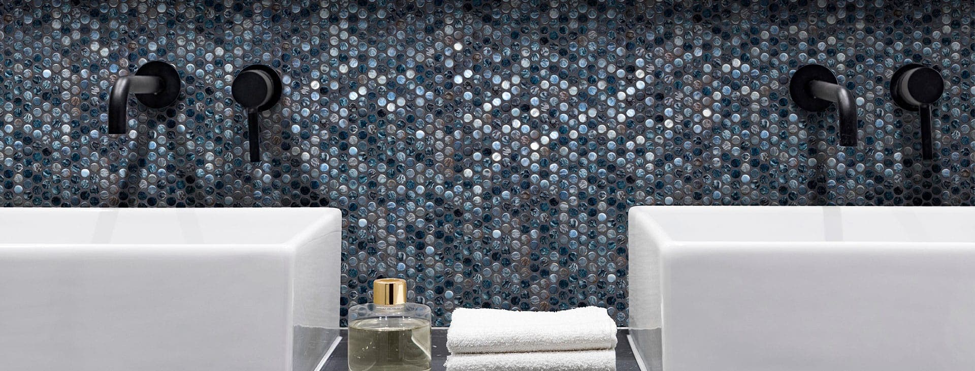 Muro de baño revestido de mosaico circular tipo penny en tonos azul, gris y negro con reflejos iridiscentes, con dos lavabos blancos, llaves negras empotradas, un difusor de vidrio y toallas blancas dobladas.