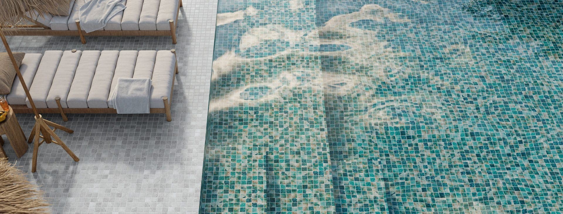 Vista desde arriba de una alberca revestida de mosaico cuadrado en tonos verde, turquesa y azul con reflejos de luz, junto a una terraza de mosaico gris claro con dos camastros de madera y colchonetas blancas.