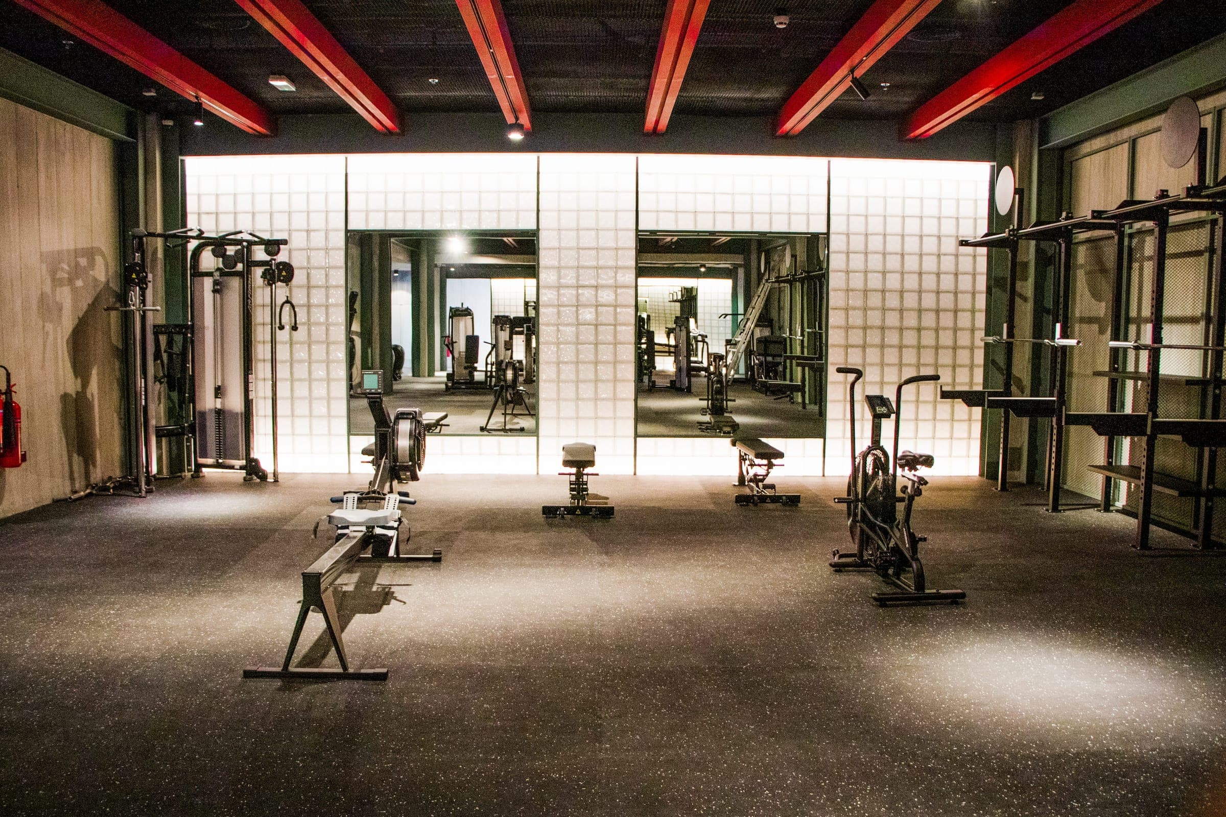 Sala de entrenamiento con piso de hule gris oscuro moteado, una remadora, una bicicleta de aire y dos bancos frente a un muro de bloques de vidrio iluminado con dos espejos.