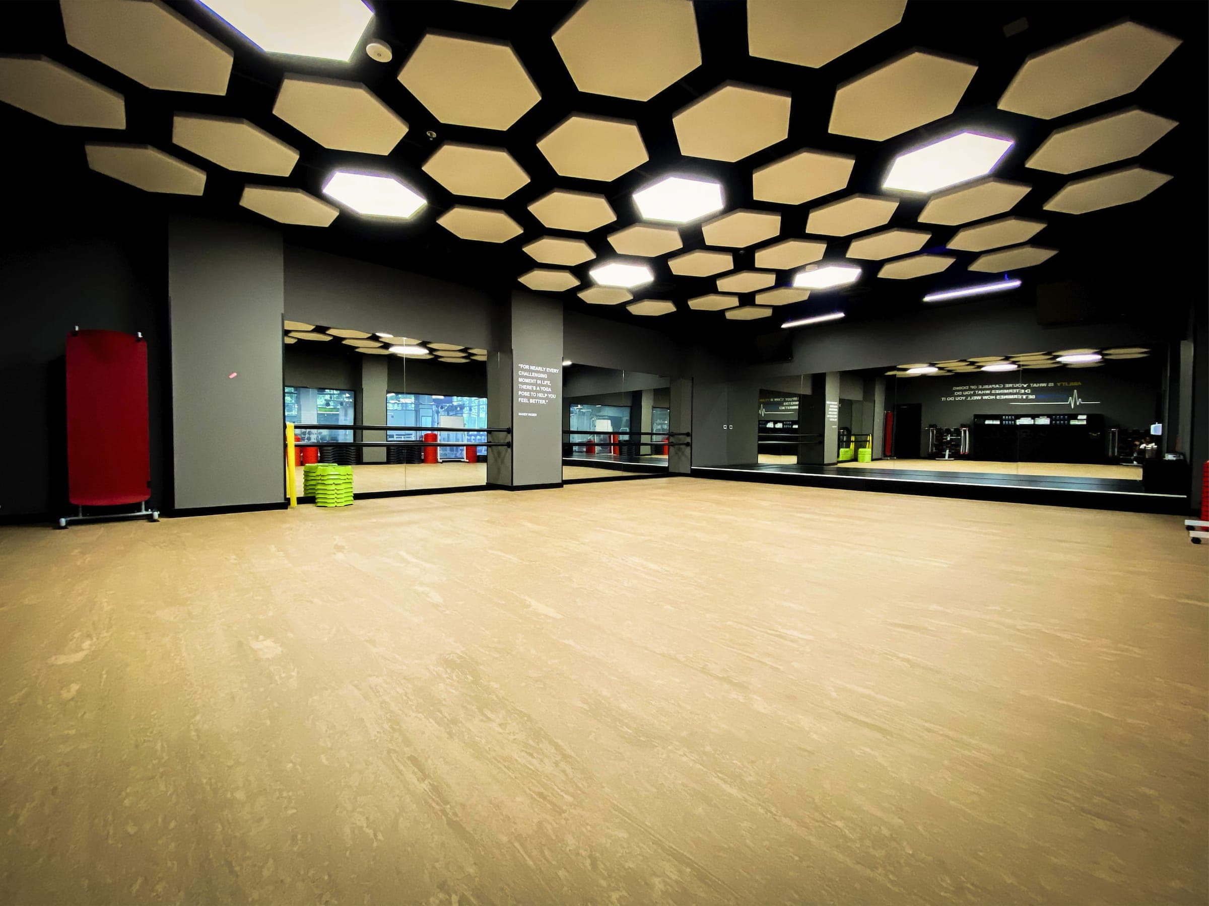 Salón de clases grupales vacío con piso de hule color haya, muros de espejos con barras, steps verdes apilados y plafón de paneles hexagonales iluminados.