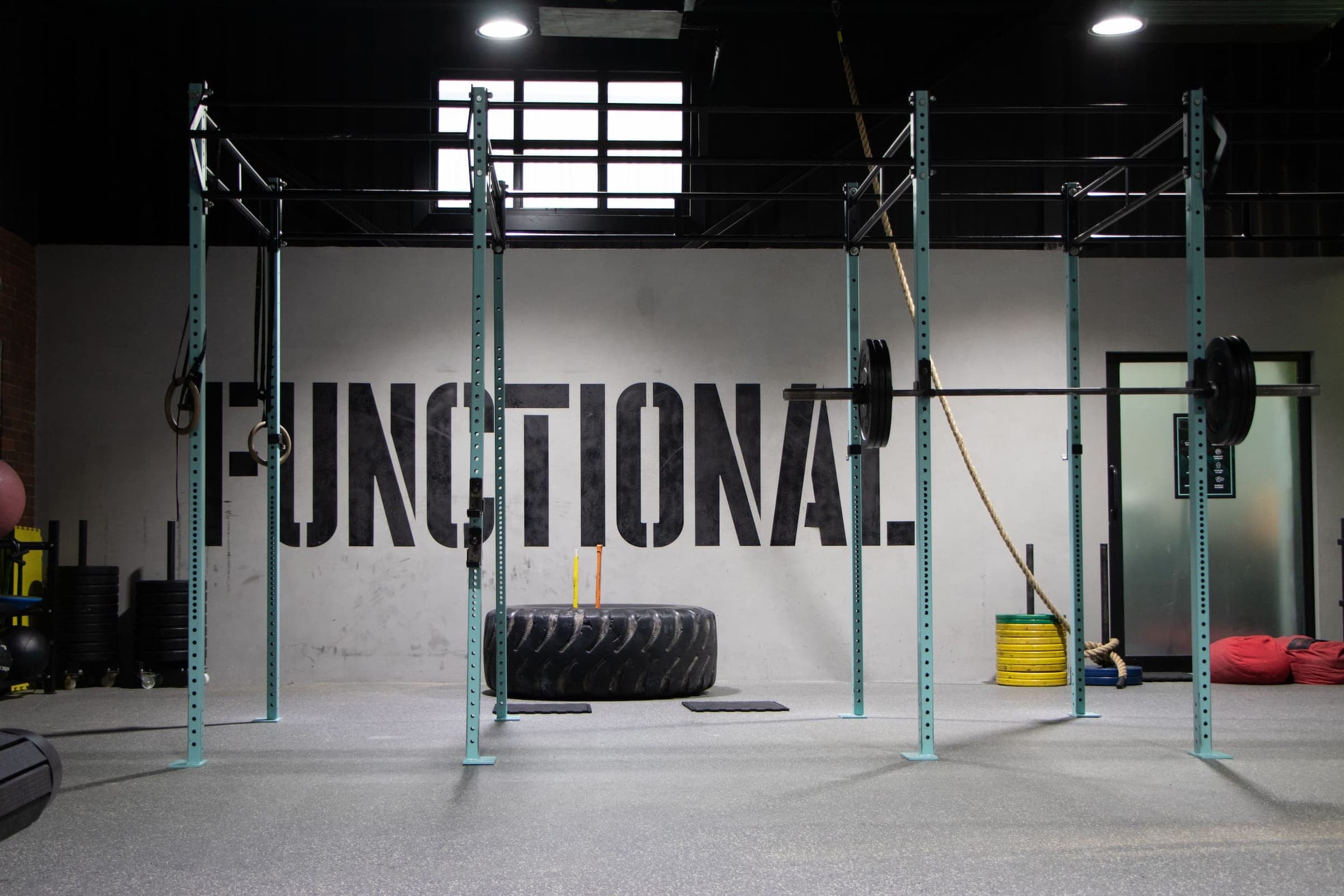 Zona de entrenamiento funcional con piso de hule gris moteado, una estructura verde menta con barra y discos, una llanta grande, una cuerda y la palabra FUNCTIONAL pintada en el muro.