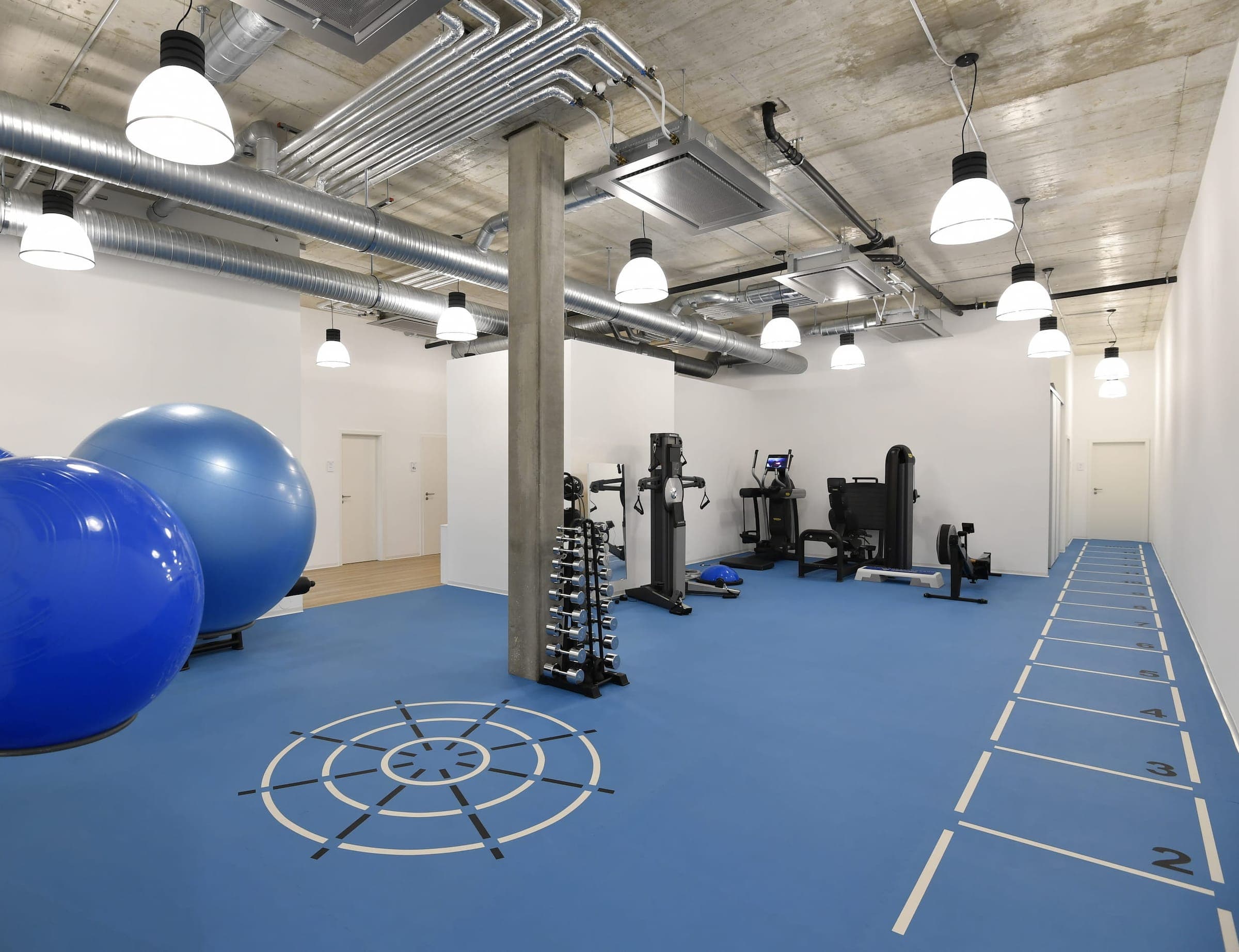 Gimnasio con piso de hule azul con un blanco y un carril numerado marcados en el piso, balones de ejercicio, una columna de concreto y máquinas al fondo.