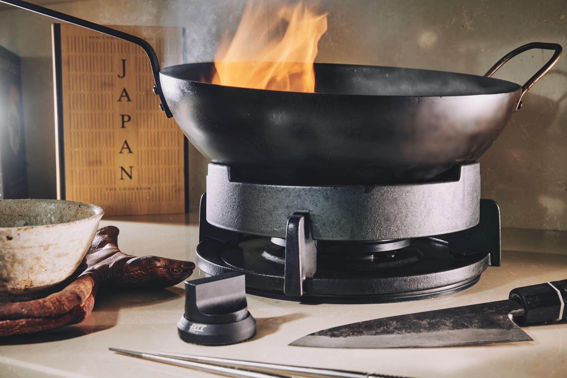Wok con flama sobre un anillo para wok montado en un quemador de parrilla negra; al frente, una perilla negra con el logotipo PITT y un cuchillo.