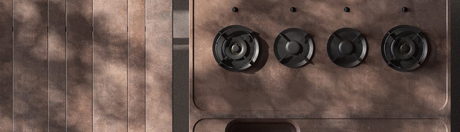 Vista cenital de una cocina exterior color óxido con cuatro quemadores de parrilla negra y cuatro perillas, junto a una mesa de listones, bajo sombras de hojas.