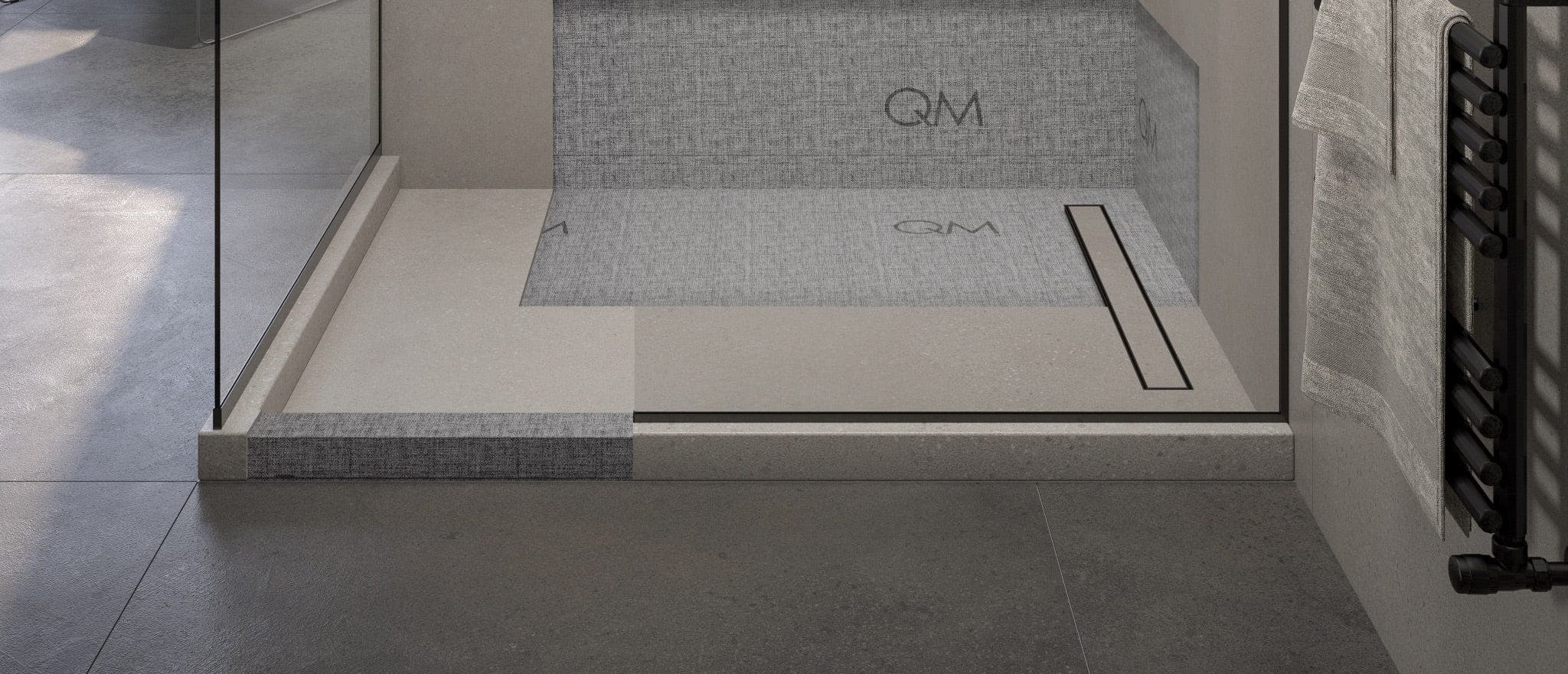 Regadera con base prefabricada de borde elevado: parte del piso y del muro muestran la membrana gris con el logotipo QM impreso y el resto ya lleva losetas; junto al muro, una coladera lineal de marco negro, y a la derecha un toallero negro con toalla.