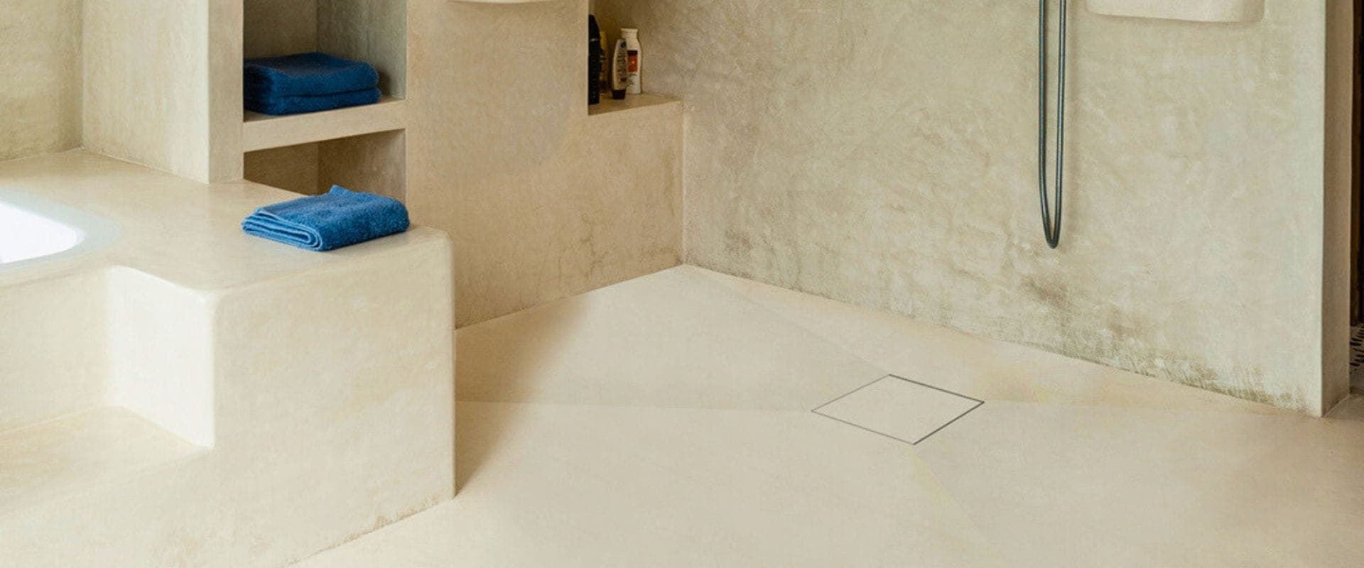 Baño con muros, tina de obra y piso continuos en tono crema, nichos con toallas azules y regadera de mano; en el piso, una coladera cuadrada cubierta con el mismo acabado, visible solo por su contorno.