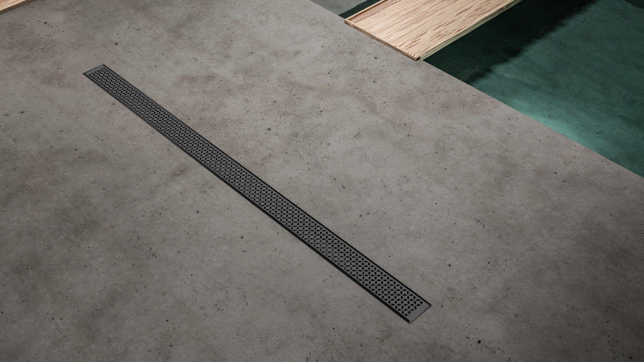 Coladera lineal negra con rejilla de perforaciones decorativas, en diagonal sobre un piso gris tipo cemento; arriba a la derecha, una tabla de madera clara sobre una superficie verde.