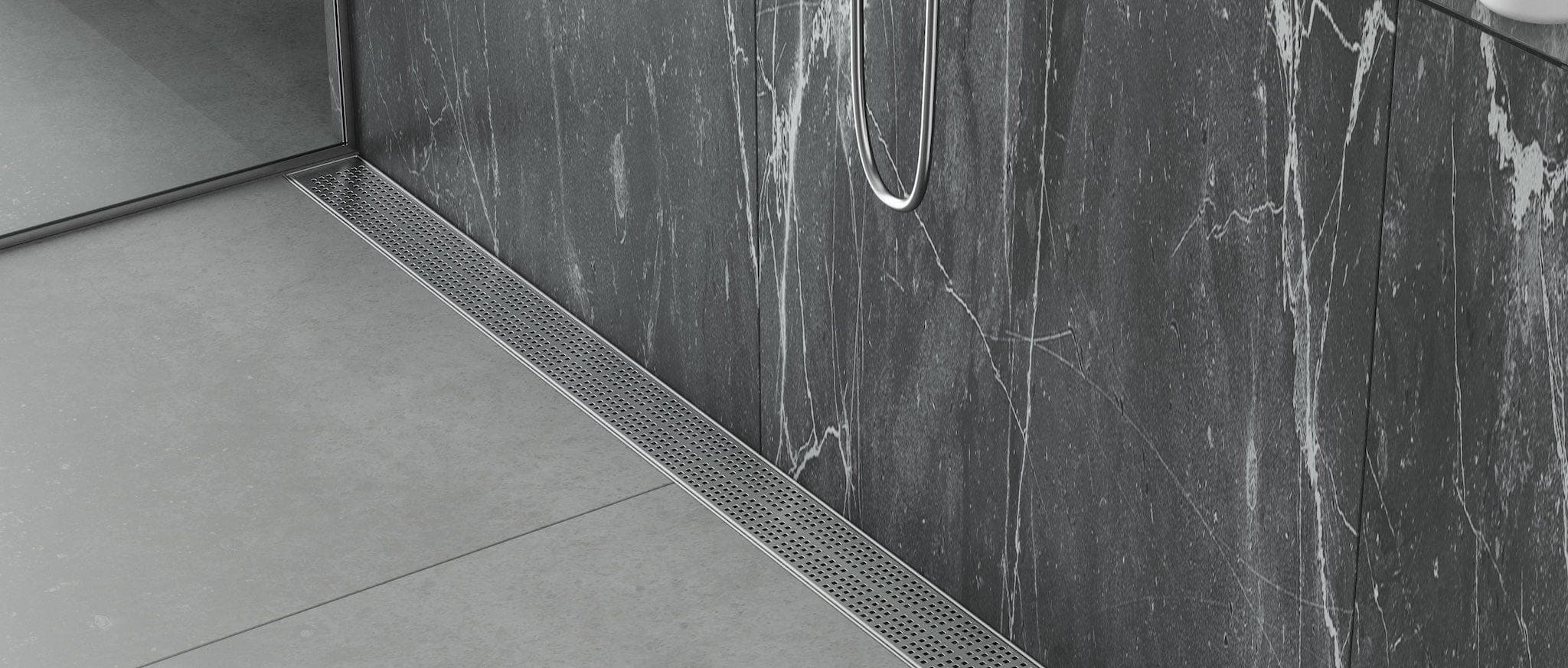 Coladera lineal de acero inoxidable con rejilla de pequeñas perforaciones cuadradas, al pie de un muro de regadera con revestimiento oscuro de vetas blancas; piso gris claro, regadera de mano y cancel de vidrio a la izquierda.