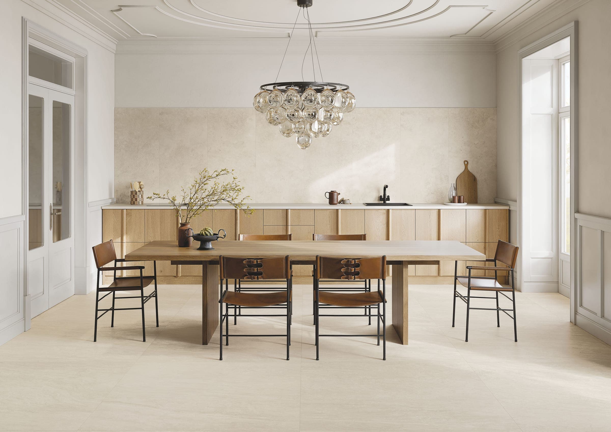 Cocina comedor con techo de molduras y una lámpara de esferas de vidrio sobre una mesa larga de madera con sillas de cuero café; el muro de la cocina está revestido con piezas grandes beige claro con aspecto de piedra y el piso es claro, con vetas finas.