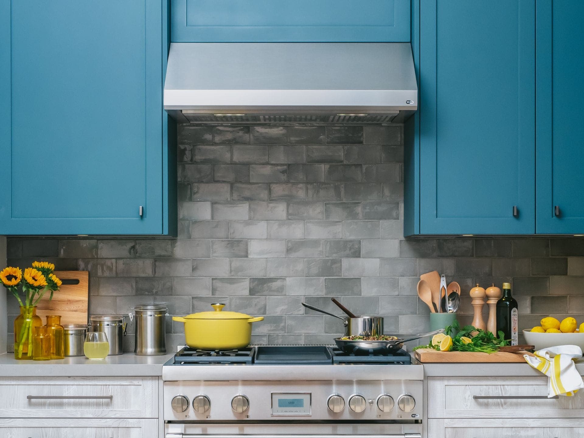 Campana de pared de acero inoxidable entre gabinetes azul turquesa, sobre un muro de azulejo gris y una estufa profesional con una olla amarilla y sartenes; a los lados, girasoles, frascos, limones y utensilios.