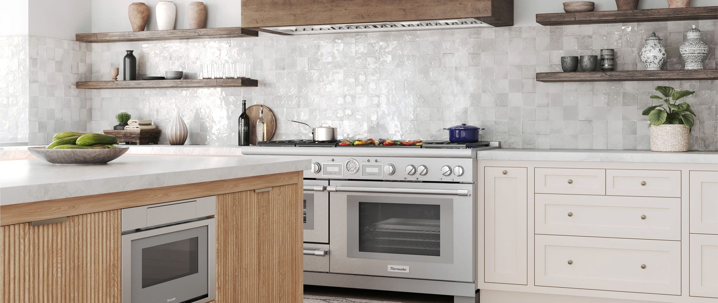 Estufa profesional de acero inoxidable con dos hornos, termómetro al centro y plancha con verduras asadas, bajo una campana empotrada en madera rústica y frente a un muro de azulejo blanco artesanal; en primer plano, una isla de madera acanalada.