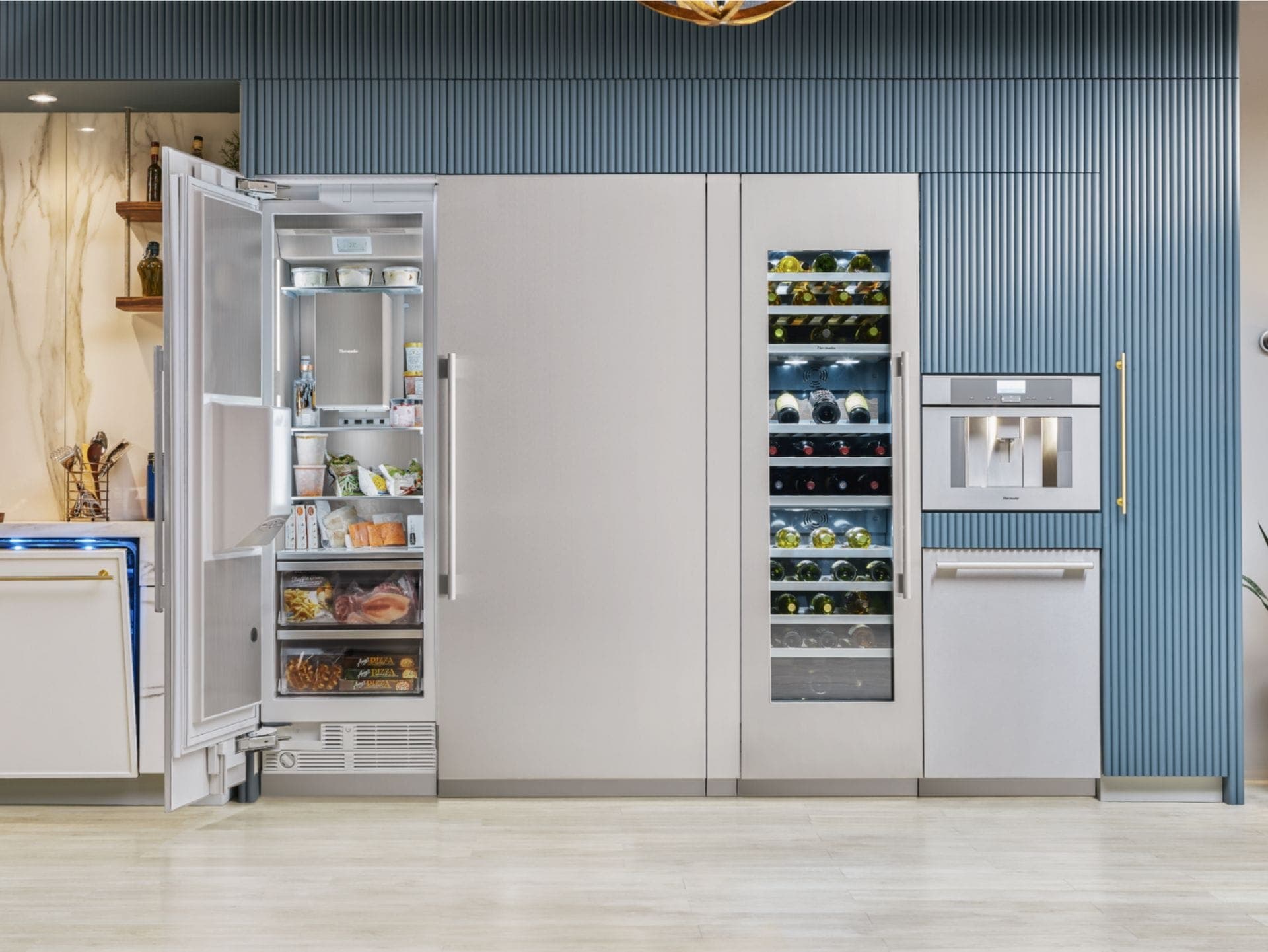 Columnas de acero inoxidable frente a un muro de listones azul: una columna abierta con alimentos congelados en repisas y cajones, otra cerrada, una cava de puerta de vidrio iluminada con botellas y una cafetera empotrada.
