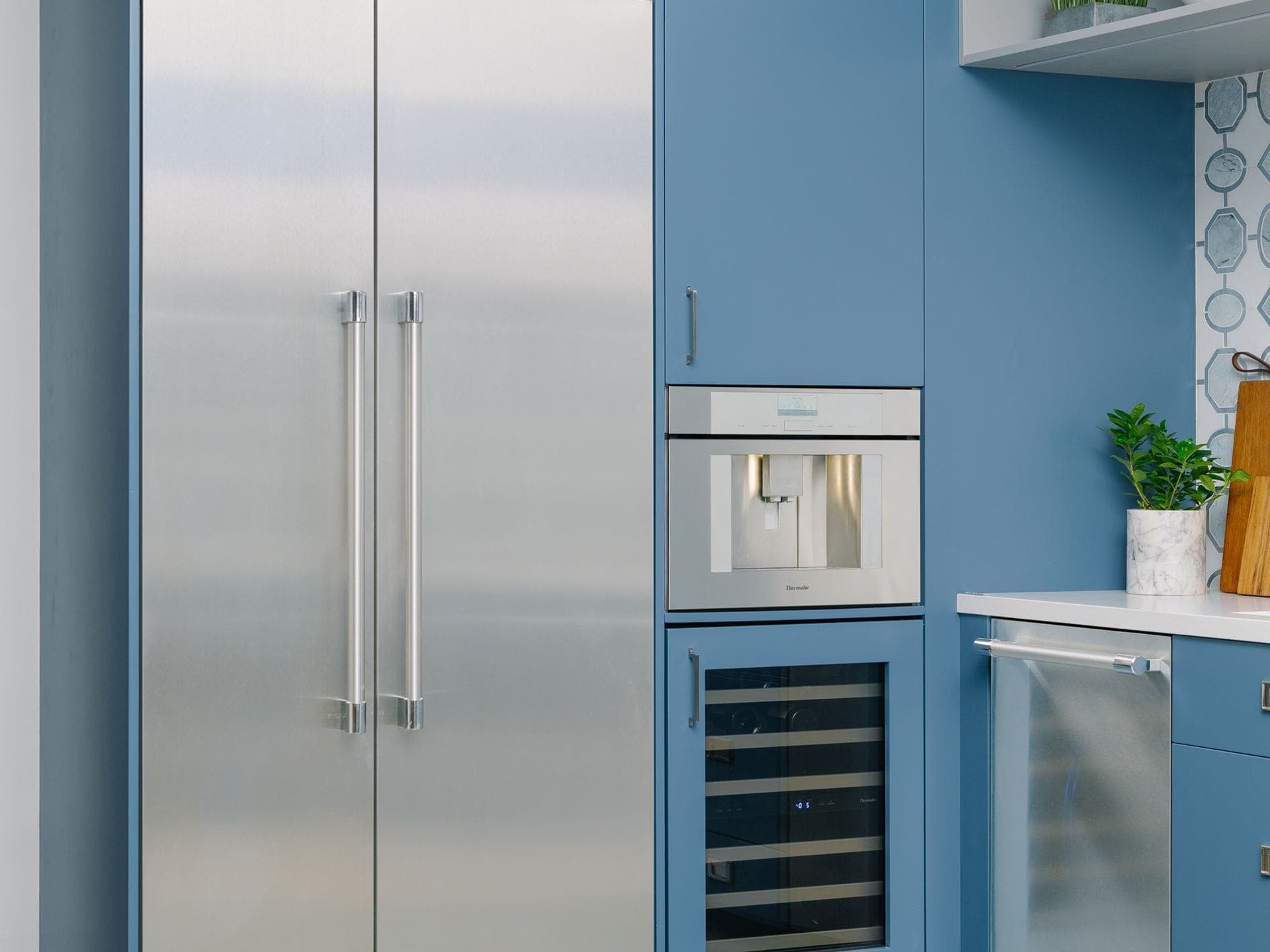 Dos columnas de acero inoxidable con jaladeras tubulares junto a una cafetera empotrada y una cava bajo cubierta con puerta de vidrio enmarcada en panel azul, en un mueble azul; a la derecha, un aparato de acero bajo la barra.