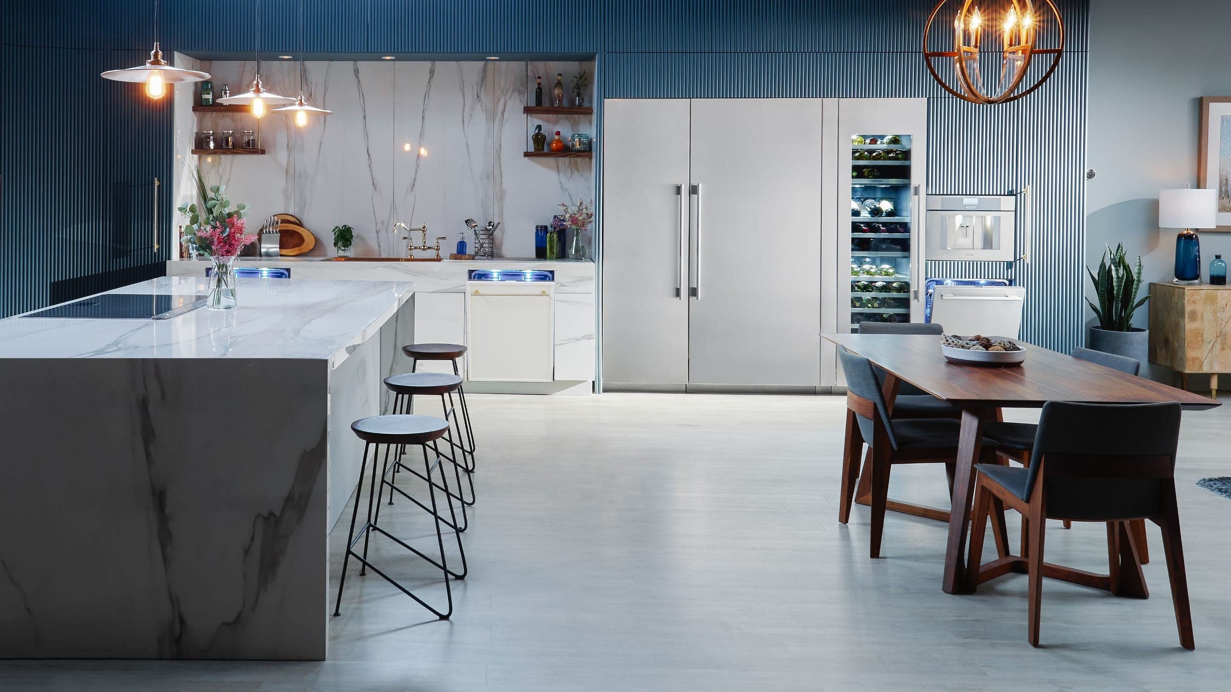 Cocina amplia con muro de listones azul oscuro: dos columnas de acero inoxidable, una cava iluminada y una cafetera empotrada; isla de mármol veteado con bancos altos, mesa de madera con sillas y lámparas colgantes.