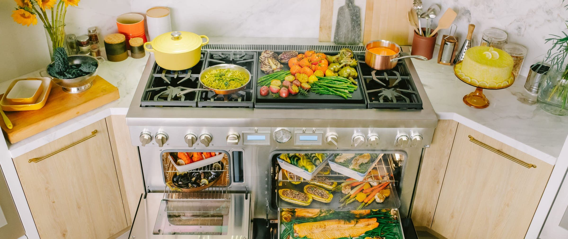 Vista superior de una estufa profesional de acero inoxidable con quemadores, plancha doble con verduras asadas, ollas y sartenes; sus dos hornos abiertos muestran charolas con mariscos, pescado, calabaza y verduras.