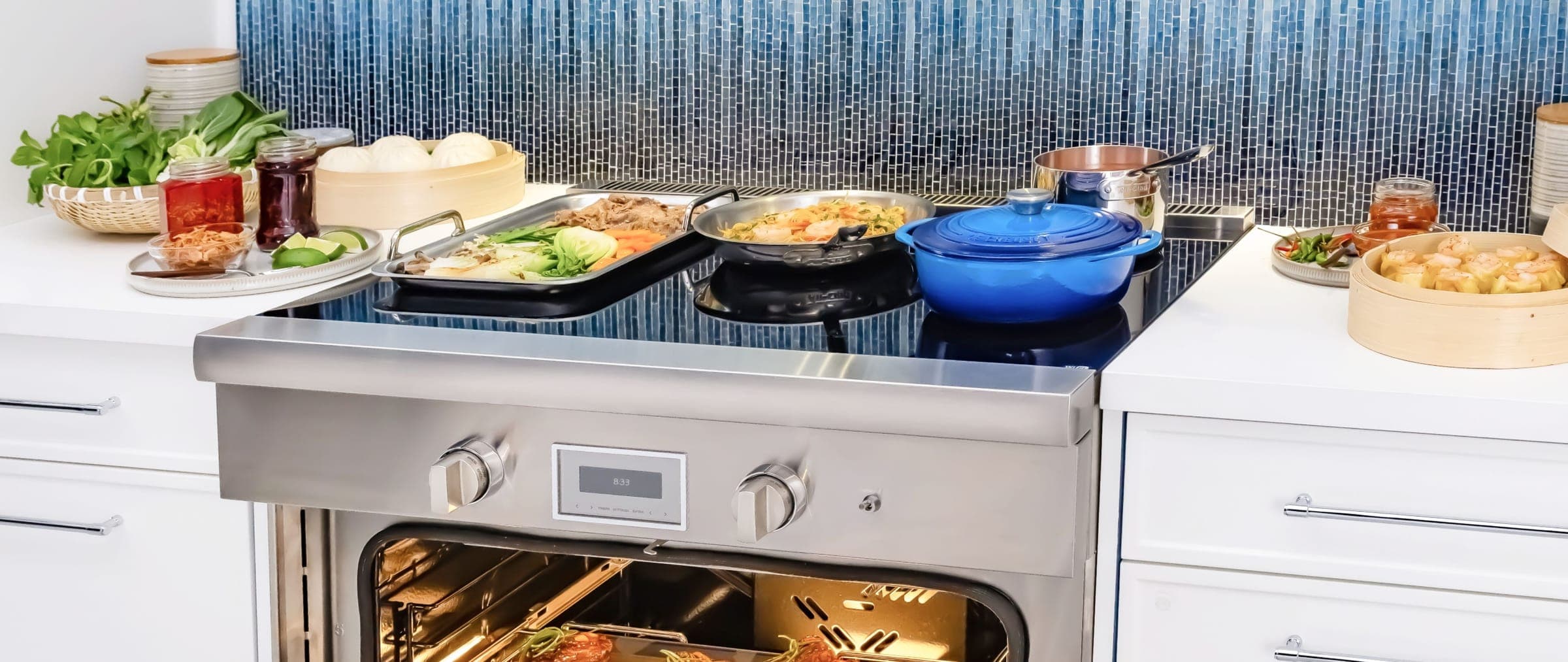 Estufa de inducción de acero inoxidable con superficie de vidrio negro, dos perillas y una pantalla en el panel; encima, una charola con verduras, un sartén, una olla azul y una cacerola; el horno está abierto e iluminado, frente a un muro de mosaico azul.