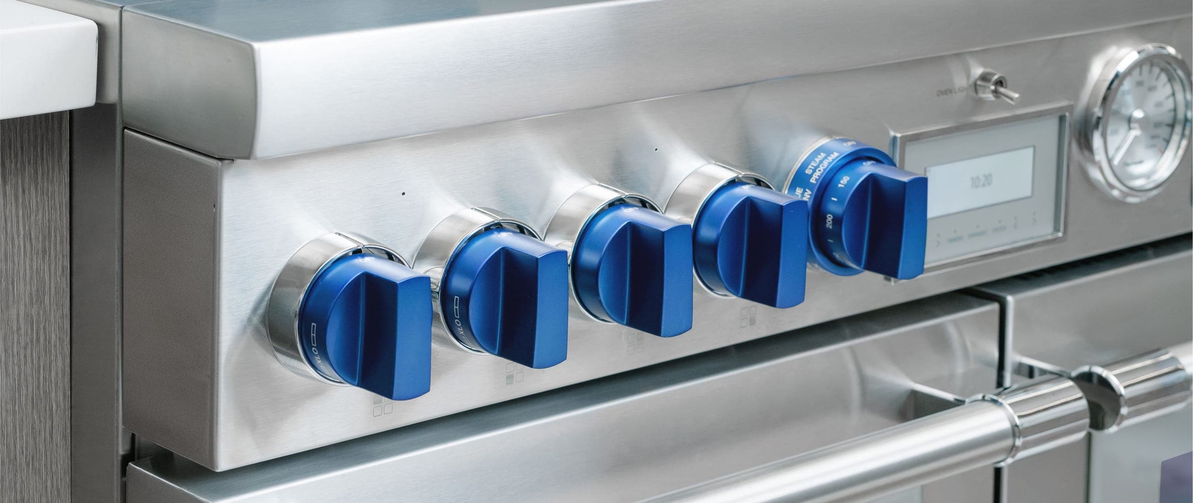 Acercamiento al panel de acero inoxidable cepillado de una estufa profesional con cinco perillas azules, una pantalla digital, un termómetro de carátula redonda y la jaladera tubular del horno.