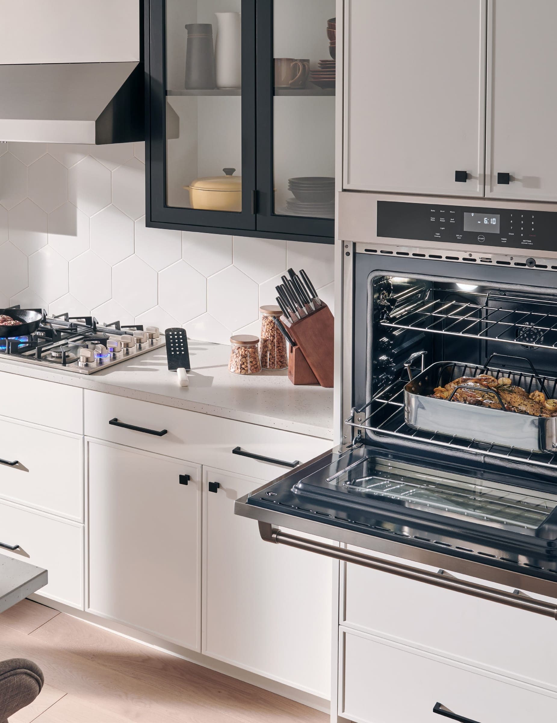 Horno de pared de acero inoxidable con la puerta abierta y una charola con comida en la rejilla, empotrado en un mueble alto blanco; a la izquierda, una cubierta de gas encendida con un sartén, gabinetes blancos con jaladeras negras, una vitrina de marco negro y un muro de azulejo hexagonal blanco.