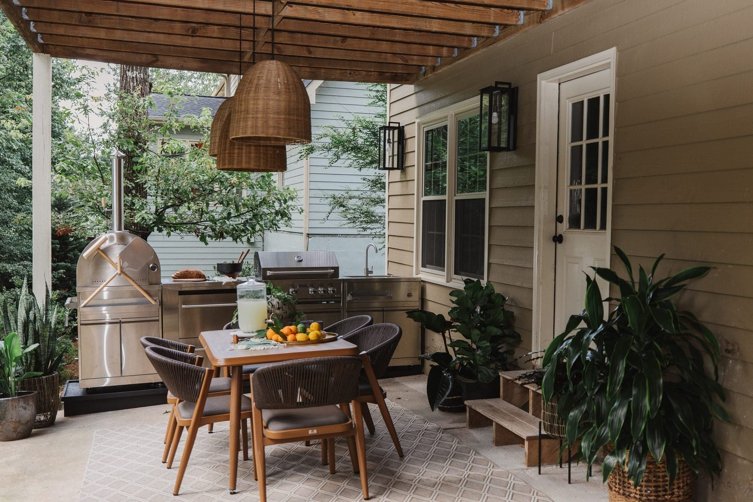 Terraza techada con pérgola de madera: módulos de acero inoxidable con horno para pizza con chimenea, gabinete, asador de gas con tapa y tarja con grifo, frente a una mesa de madera con sillas tejidas, dos lámparas colgantes de mimbre y plantas en macetas.