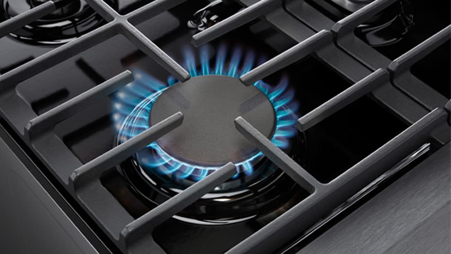 Acercamiento a un quemador de gas encendido, con un anillo de flama azul bajo una tapa negra y parrillas negras alrededor.