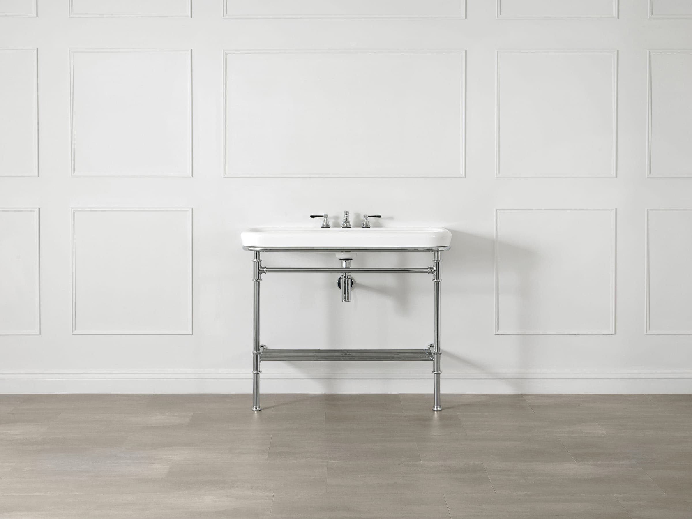 Consola de cromo con lavabo rectangular blanco, mezclador de tres piezas, travesaño y repisa de rieles, frente a un muro blanco con molduras