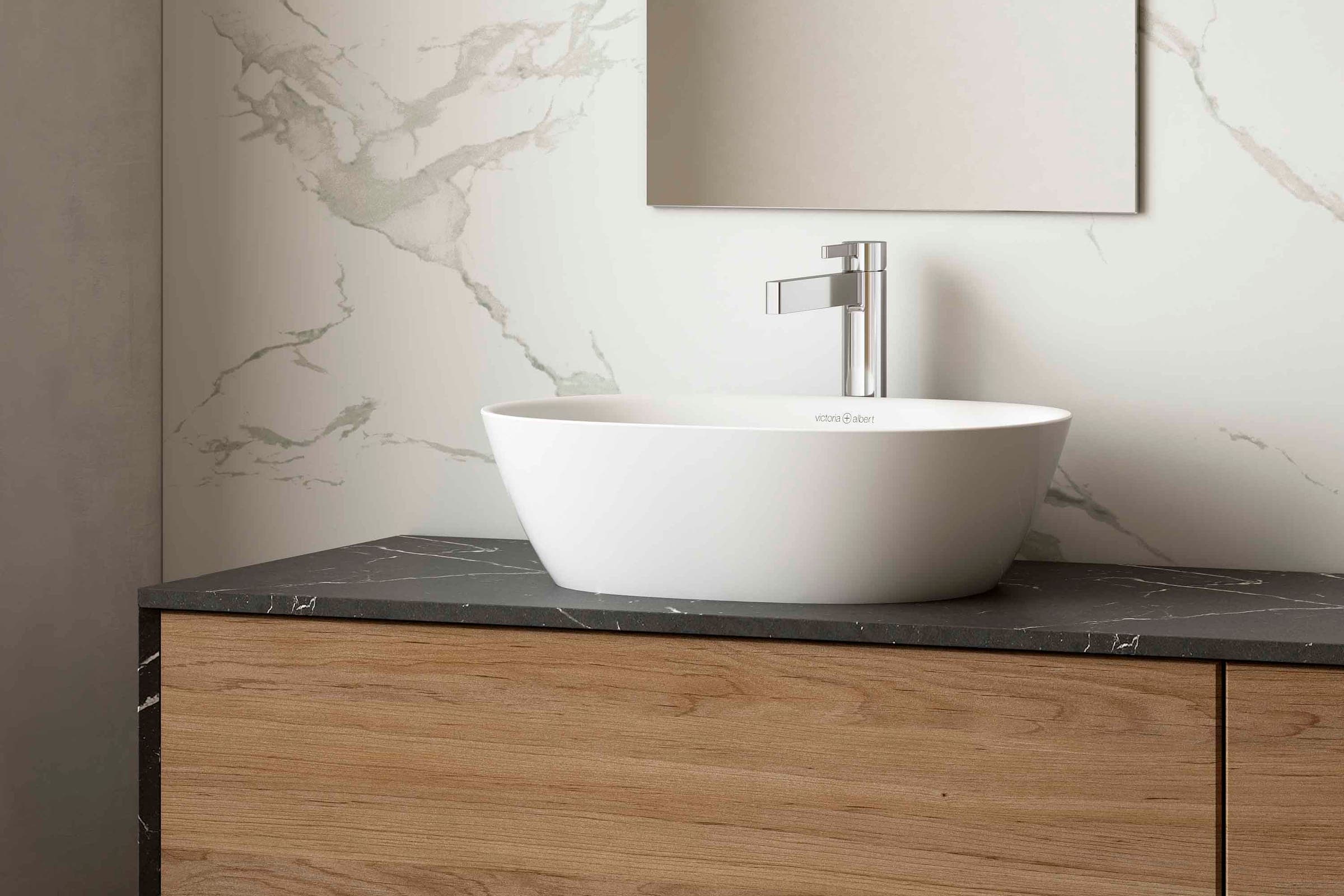 Lavabo ovalado blanco de sobreponer sobre una cubierta de piedra oscura, en un mueble de madera, con mezclador alto cromado y un espejo; muro de mármol blanco veteado