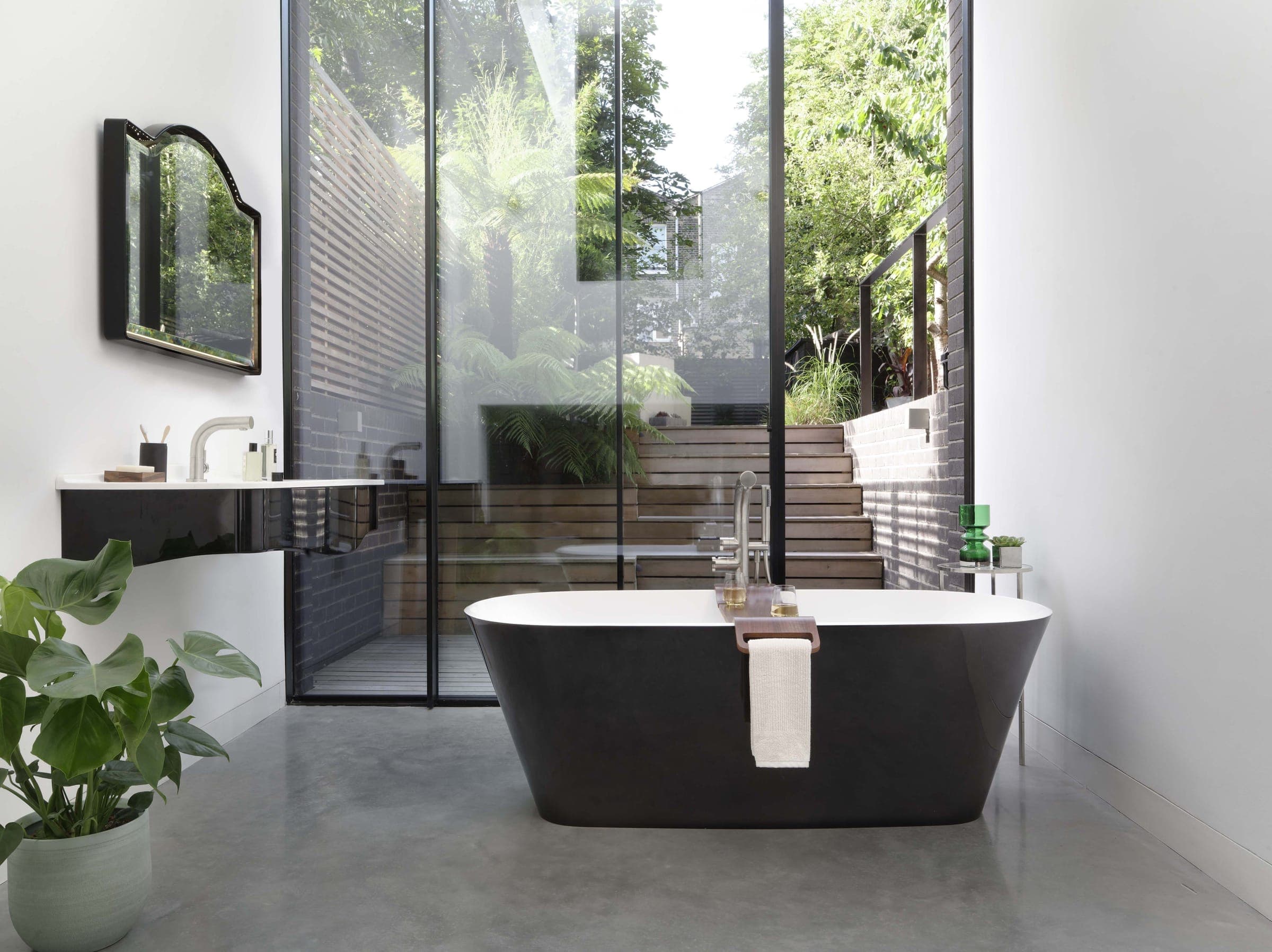 Tina exenta con exterior negro brillante e interior blanco, con una repisa de madera y una toalla, frente a un ventanal hacia un patio con helechos; a la izquierda, un lavabo de muro con exterior negro