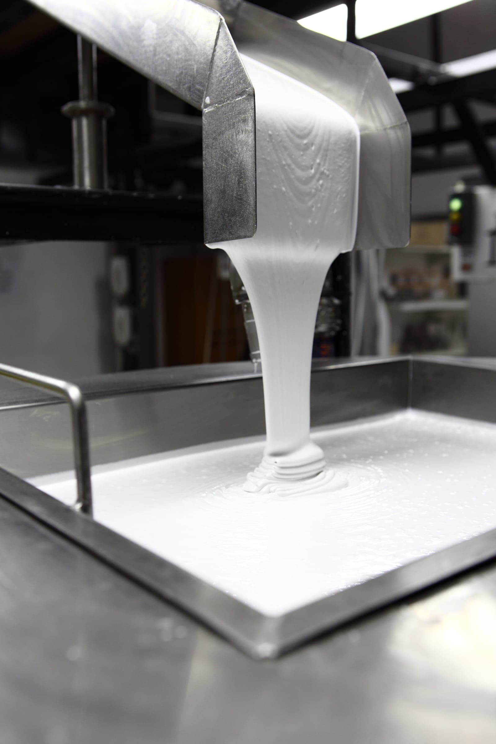 Material blanco y espeso que cae de una tolva de acero inoxidable sobre una charola metálica, en un taller