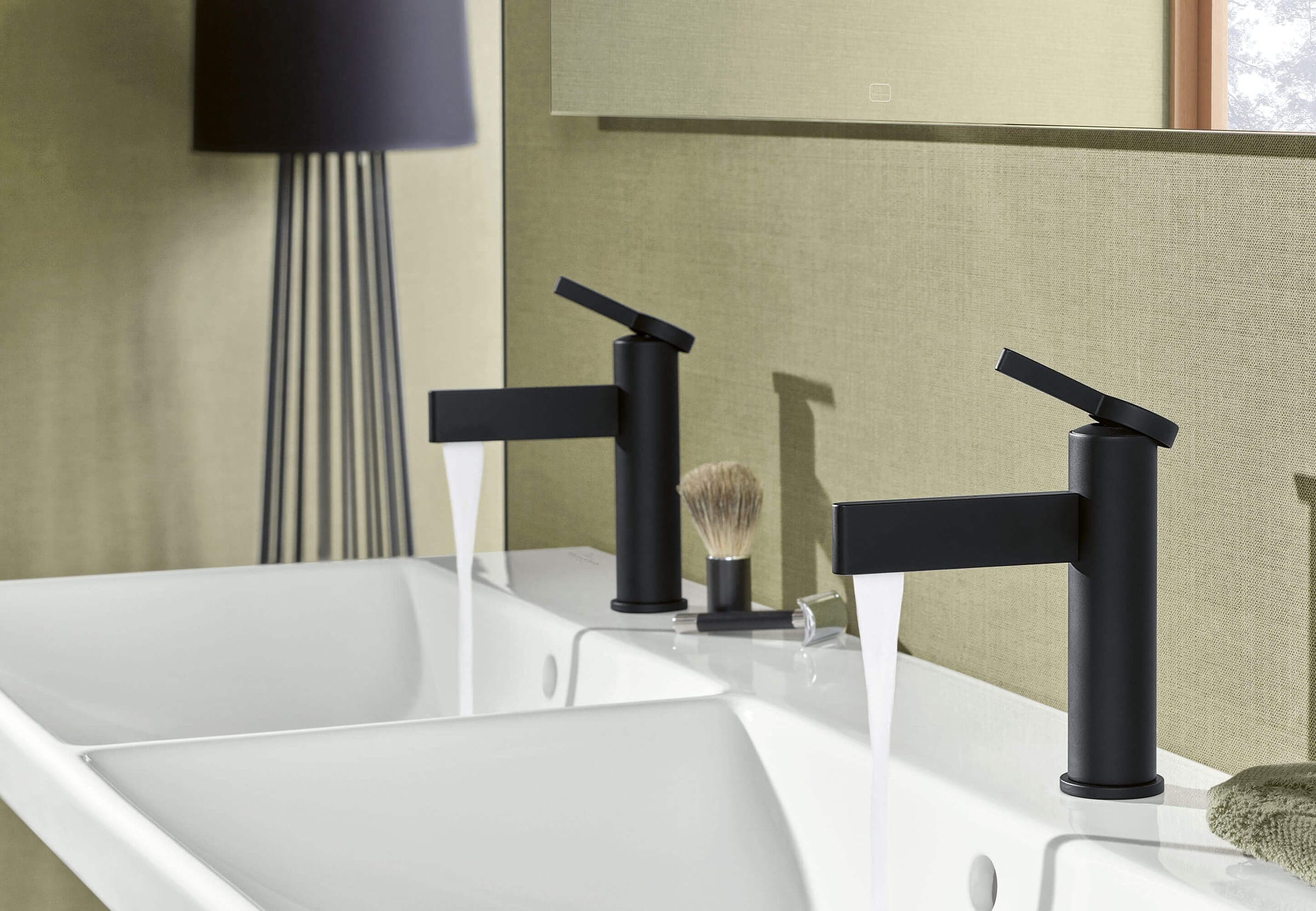 Dos monomandos negros de cuerpo cilíndrico y caño recto sobre un lavabo doble blanco, con agua corriendo; entre ellos, una brocha de afeitar; al fondo, una lámpara negra y una pared verde olivo texturizada.