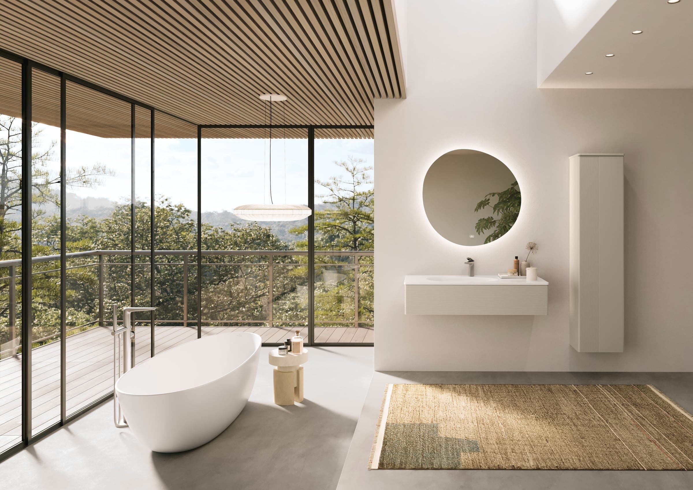 Baño amplio con ventanales de piso a techo hacia un bosque: tina exenta blanca de forma ovalada con grifería de piso, mueble de lavabo suspendido blanco de frente acanalado, espejo iluminado de contorno orgánico, mueble alto y tapete de fibra natural.