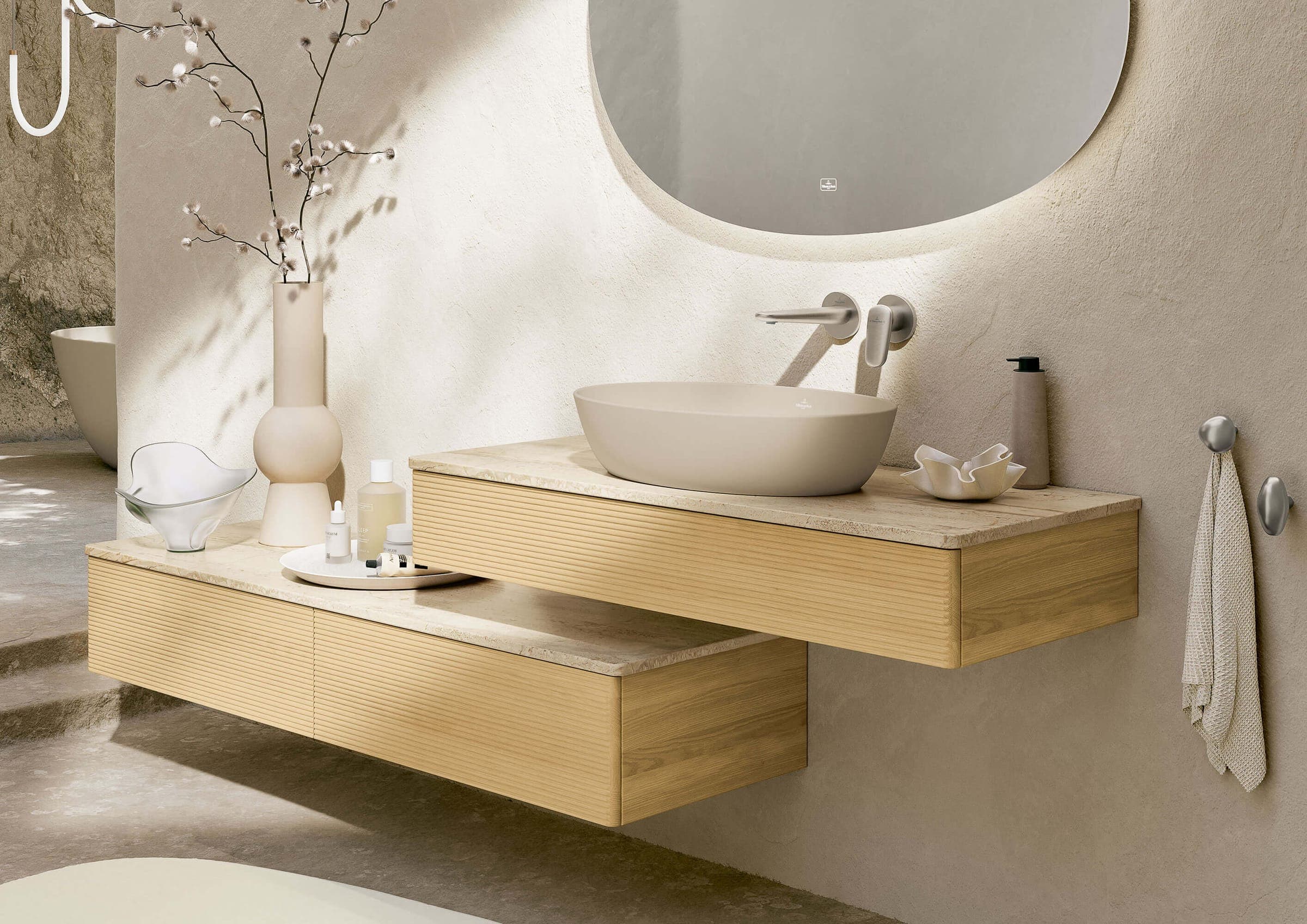 Lavabo ovalado sobre cubierta en tono almendra mate sobre dos muebles suspendidos de madera clara escalonados, con grifería de pared, espejo ovalado, un florero con ramas y una toalla colgada de un gancho.