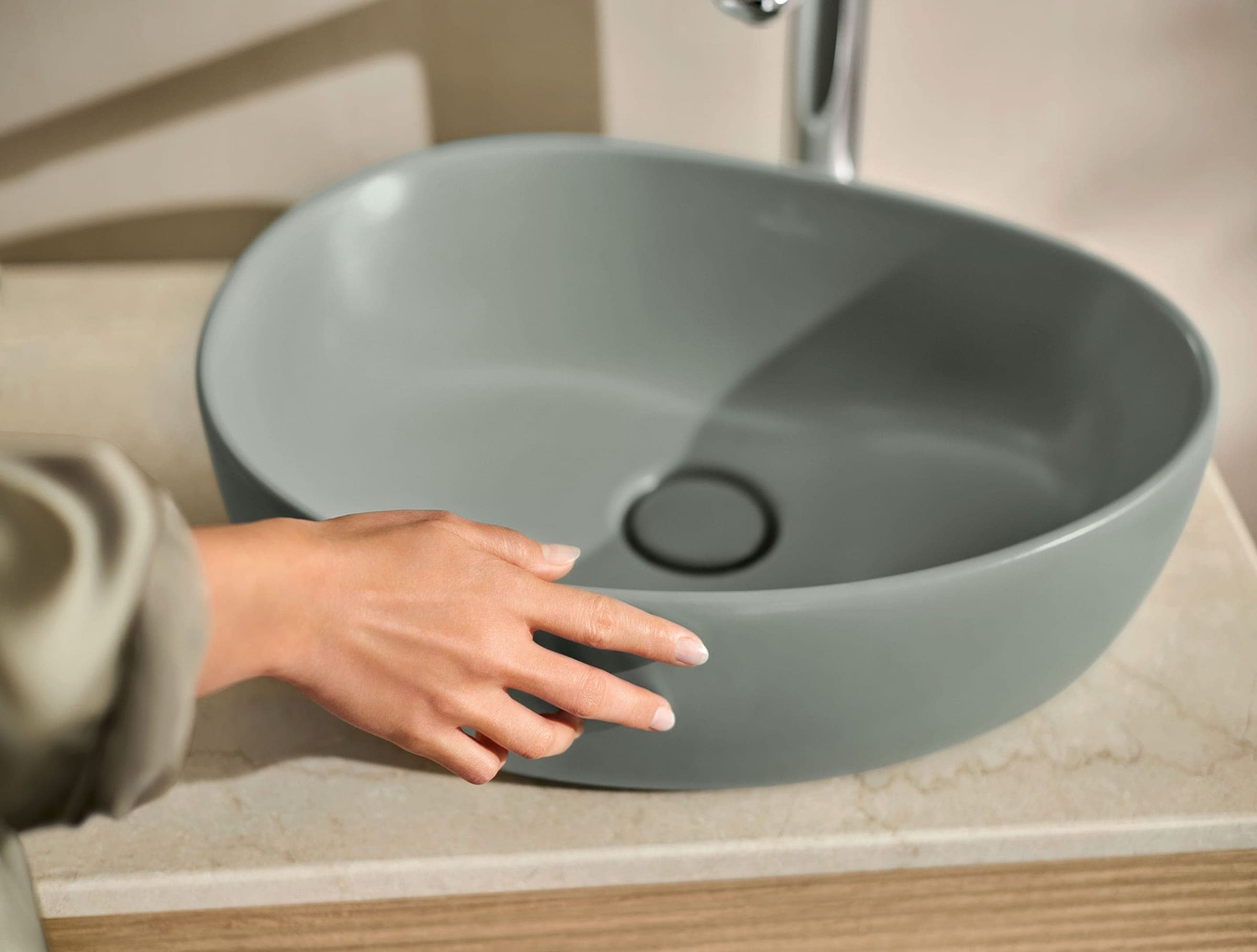 Mano que toca el borde de un lavabo verde grisáceo, mate y de forma orgánica, colocado sobre una cubierta de piedra beige; arriba asoma la base de un grifo cromado.