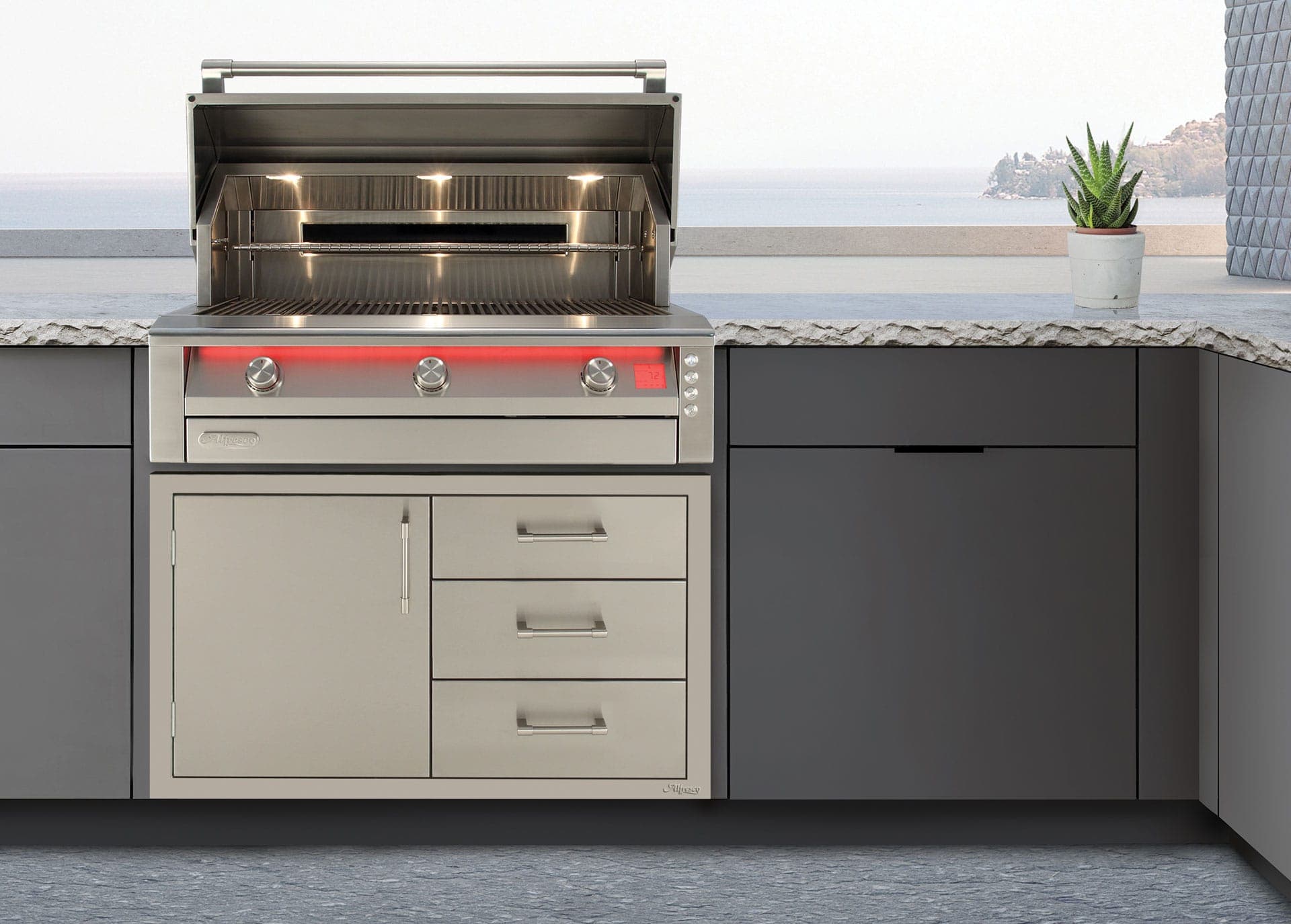 Asador de acero inoxidable con la tapa abierta y tres luces encendidas en el interior; el tablero tiene una franja de luz roja, tres perillas y una pantalla digital. Debajo, un mueble de una puerta y tres cajones, entre gabinetes gris oscuro con cubierta de piedra; al fondo, el mar y una isla.