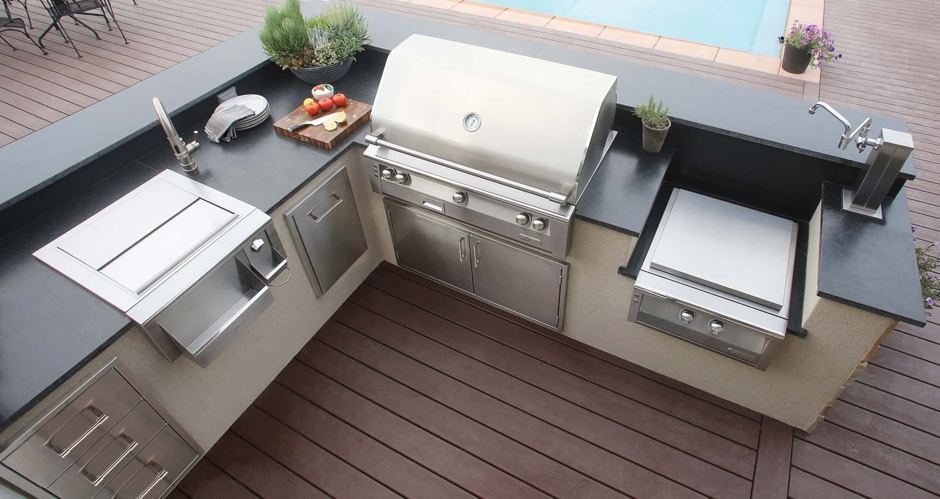 Vista desde arriba de una cocina exterior con cubierta negra junto a una alberca: asador de acero inoxidable con la tapa cerrada y termómetro sobre una puerta doble, tarja con cubierta deslizable, puerta de acceso, cajonera, un módulo de cocción tapado y dos llaves de agua; sobre la cubierta, una tabla con jitomates y limones, platos y macetas; piso de duela.