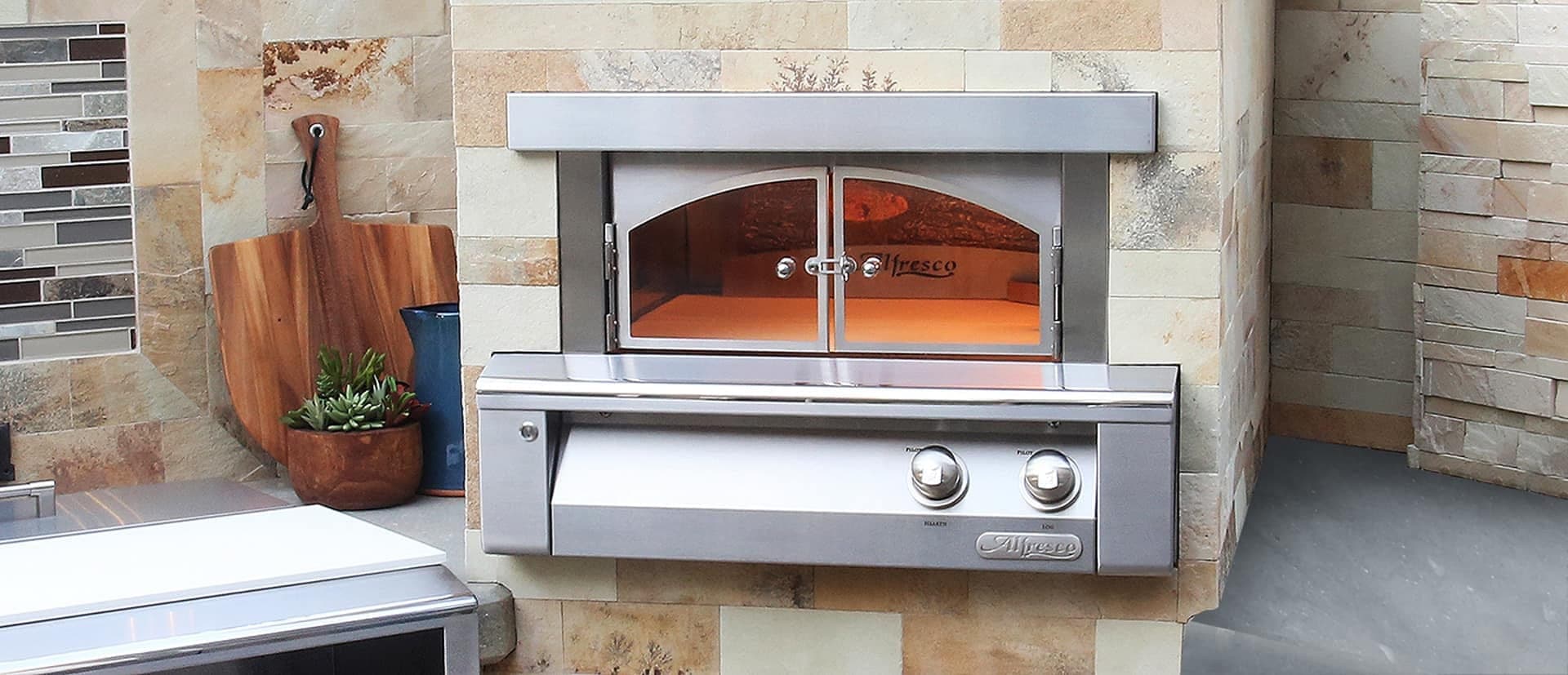 Horno para pizza de acero inoxidable empotrado en un muro de piedra, con puertas de vidrio en arco, interior iluminado y dos perillas; a la izquierda, una tabla de madera colgada, una jarra azul y suculentas en una maceta de madera, junto a un mosaico de vidrio.