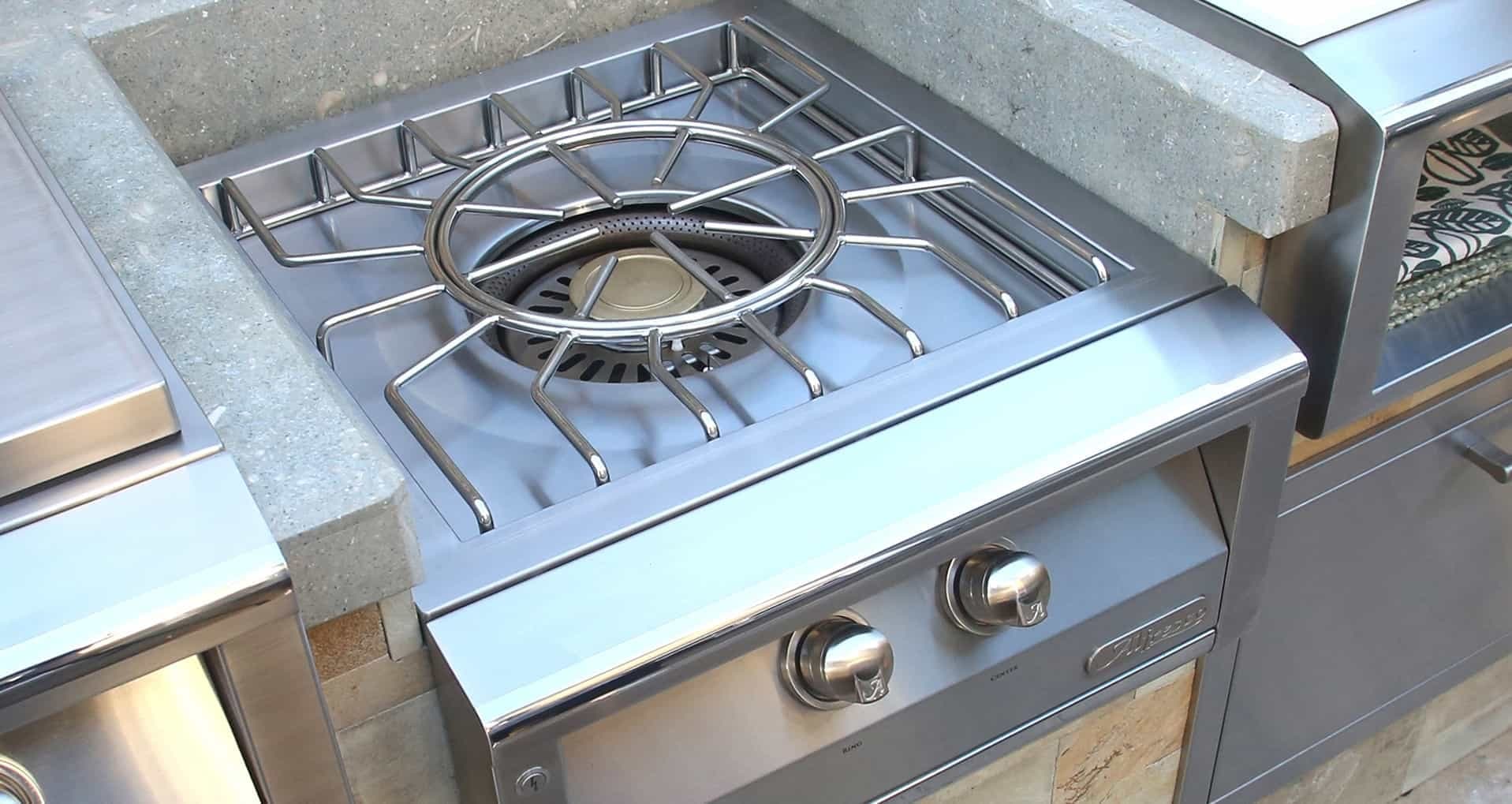 Módulo Versa Power empotrado en una cubierta de concreto: rejilla de araña de acero inoxidable con aro central, quemador de centro dorado rodeado de un anillo perforado oscuro y dos perillas metálicas con el logotipo Alfresco al frente; a los lados, otros módulos de acero inoxidable.