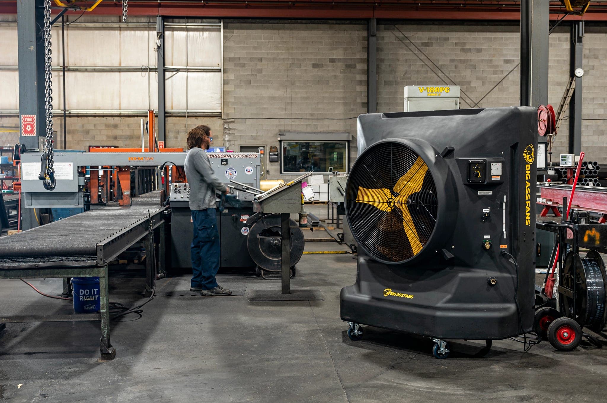 Taller metalúrgico con un transportador de rodillos y un operario frente a una máquina; a la derecha, un enfriador evaporativo negro sobre ruedas, con aspas amarillas tras una rejilla y el logotipo BIG ASS FANS impreso en el equipo.