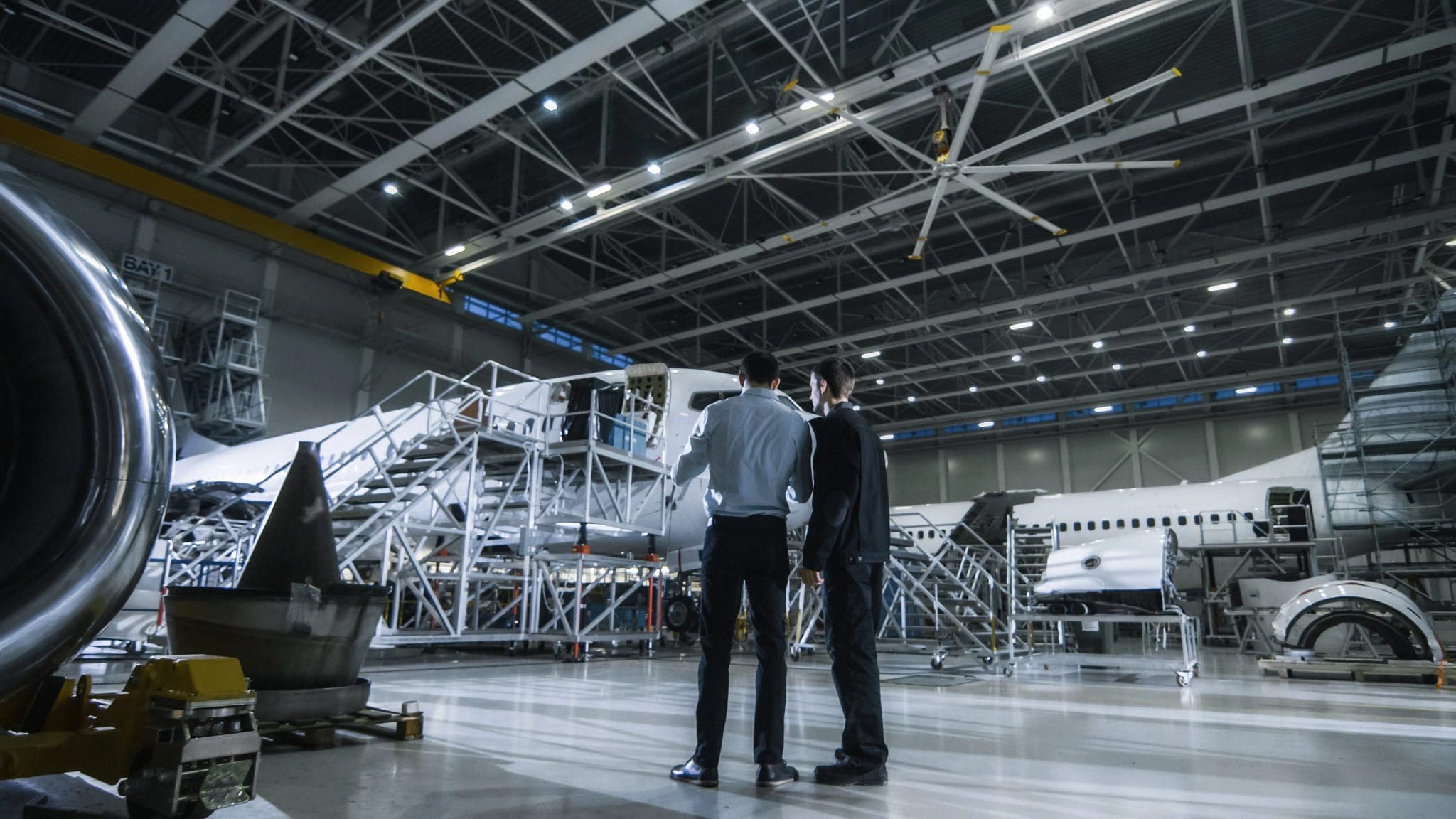 Hangar de estructura metálica con aviones en mantenimiento rodeados de andamios y escaleras; dos hombres de espaldas los observan y, en lo alto, cuelga un ventilador de techo de gran diámetro con aspas plateadas de puntas amarillas; a la izquierda, la turbina de un avión.