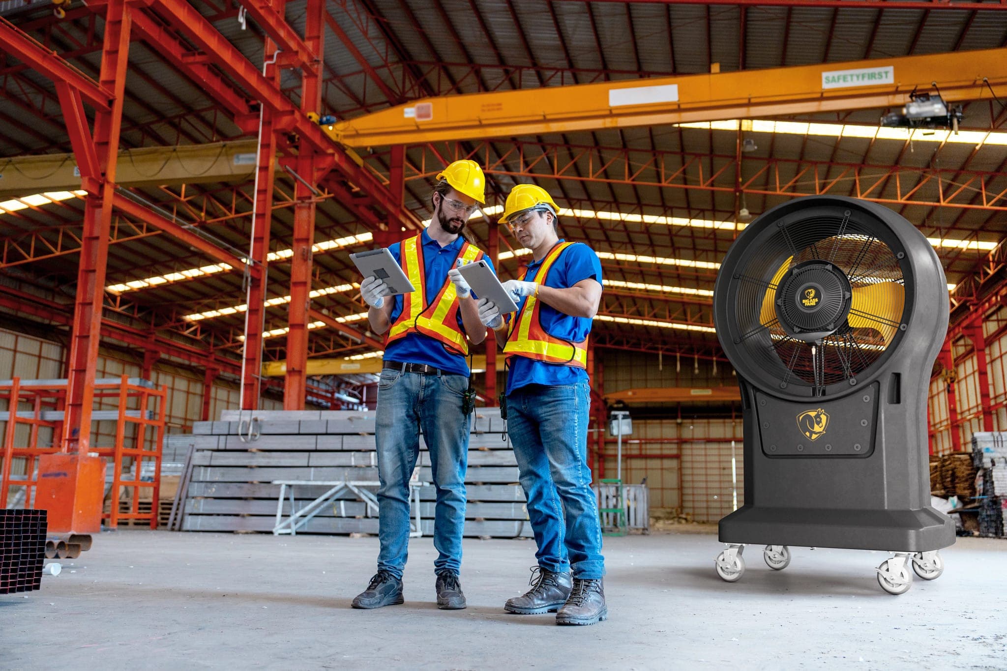 Nave industrial con estructura de acero roja y una grúa viajera amarilla; dos trabajadores con casco amarillo y chaleco reflejante revisan tabletas junto a un ventilador nebulizador negro sobre ruedas, con aspas amarillas y el logotipo de la marca al centro.