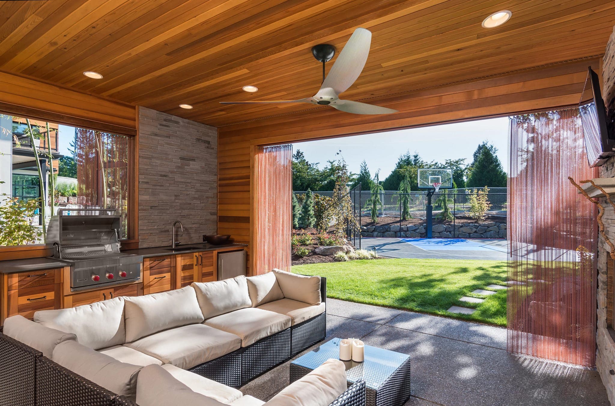 Terraza techada con plafón de madera, cocina exterior con asador de acero inoxidable y tarja, cortinas de malla metálica cobriza y una sala modular de mimbre oscuro con cojines color crema; en el techo, un ventilador de tres aspas plateadas; al fondo, jardín y una cancha de basquetbol.